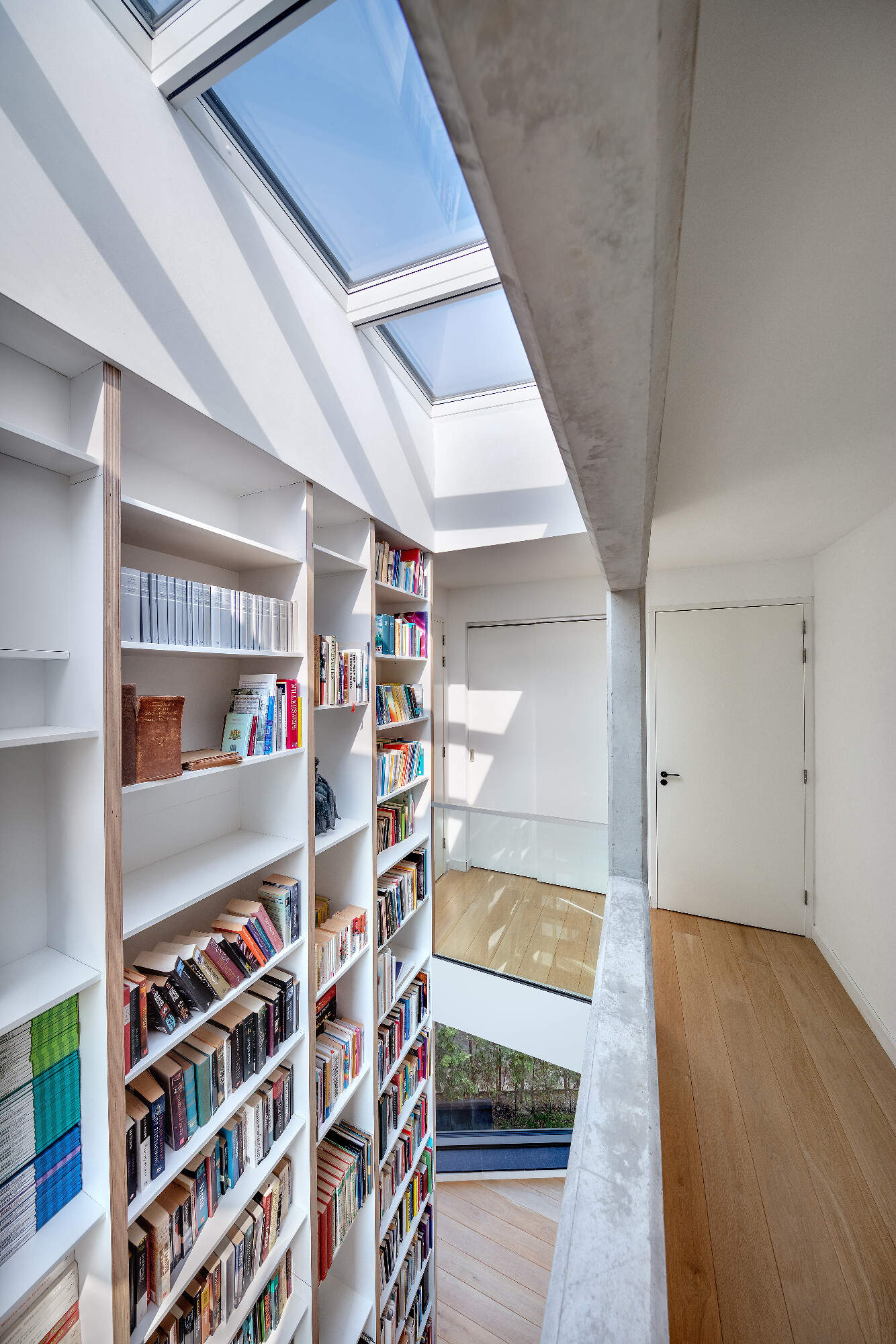 A central staircase with wood treads winds upward beside a full height custom bookshelf. Natural light from skylights and large windows keeps the vertical core bright and airy.