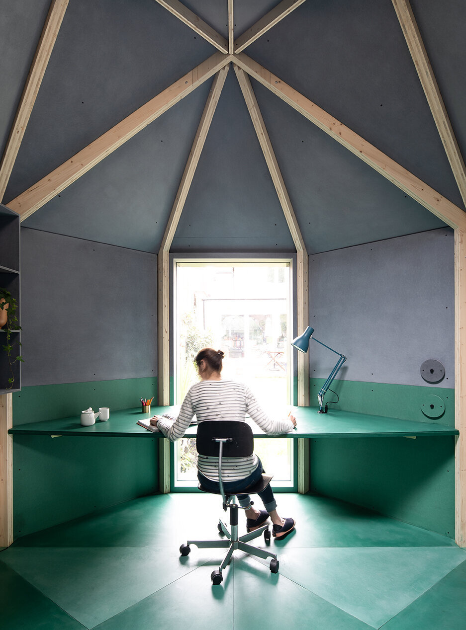 Inside this backyard studio, exposed timber framing converges at the ceiling, creating a sense of height, while angled walls, a built-in desk, and a fold-out bed make the space flexible for work, rest, or quiet contemplation.