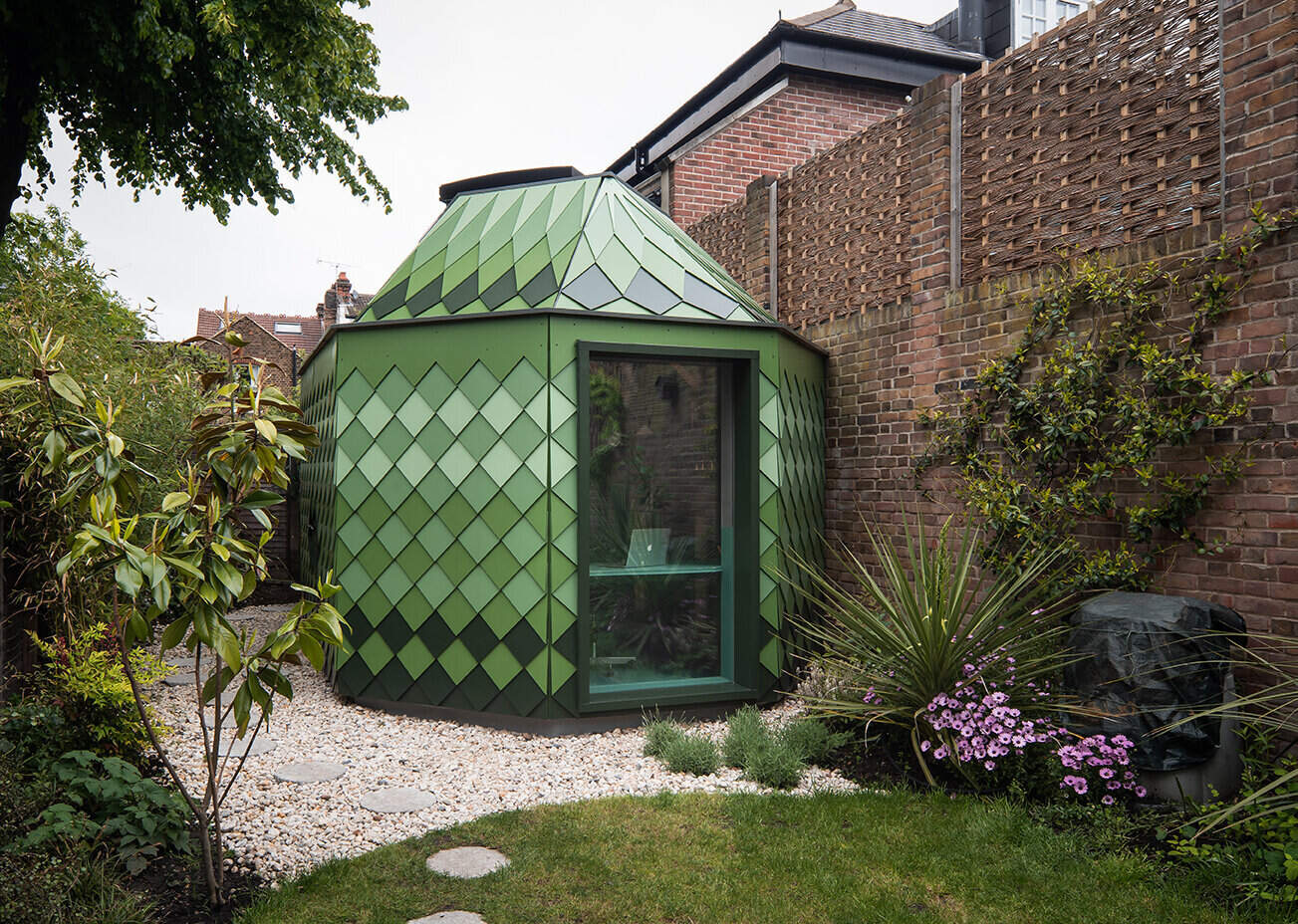 A small green-shingled studio sits tucked in a South West London backyard, its octagonal walls and hexagonal roof blending geometry with garden surroundings while hinting at the playful space within.