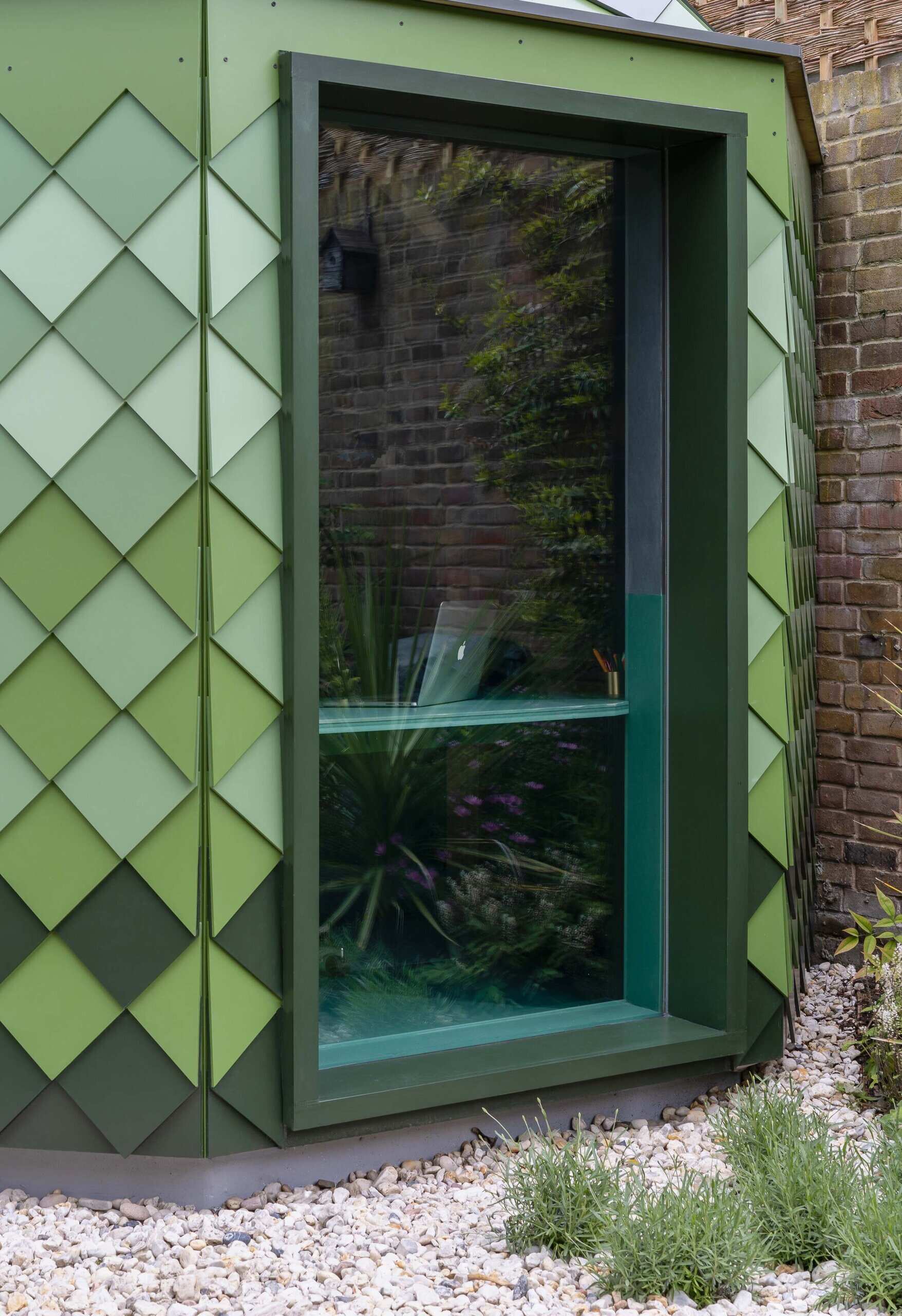 A small green-shingled studio sits tucked in a South West London backyard, its octagonal walls and hexagonal roof blending geometry with garden surroundings while hinting at the playful space within.