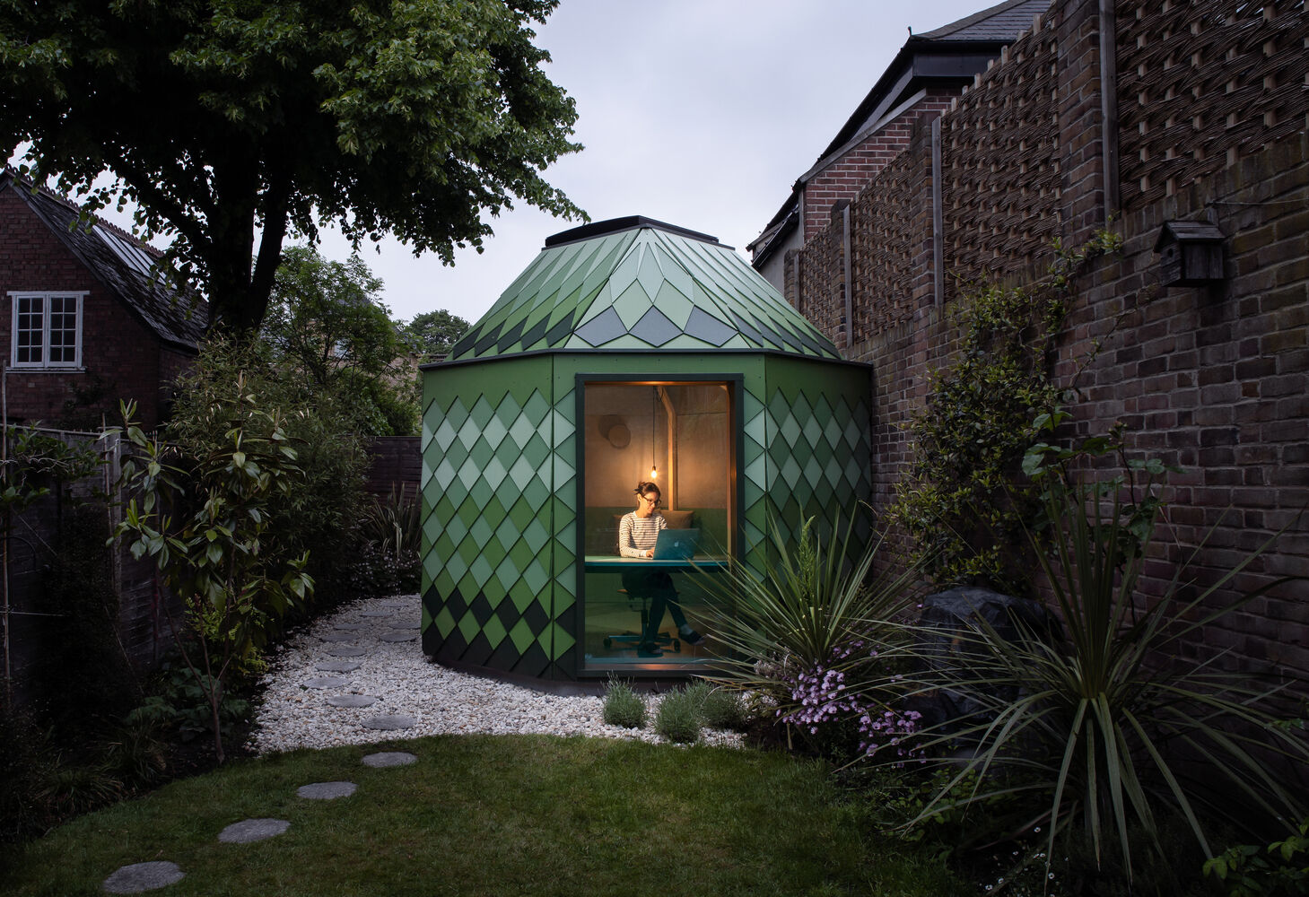 A small green-shingled studio sits tucked in a South West London backyard, its octagonal walls and hexagonal roof blending geometry with garden surroundings while hinting at the playful space within.