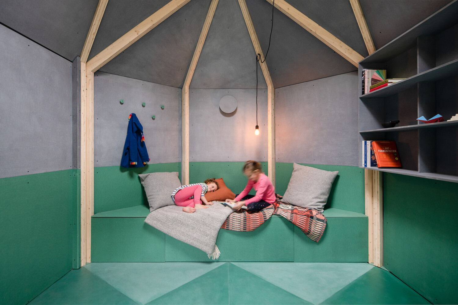 Inside this backyard studio, exposed timber framing converges at the ceiling, creating a sense of height, while angled walls, a built-in desk, and a fold-out bed make the space flexible for work, rest, or quiet contemplation.