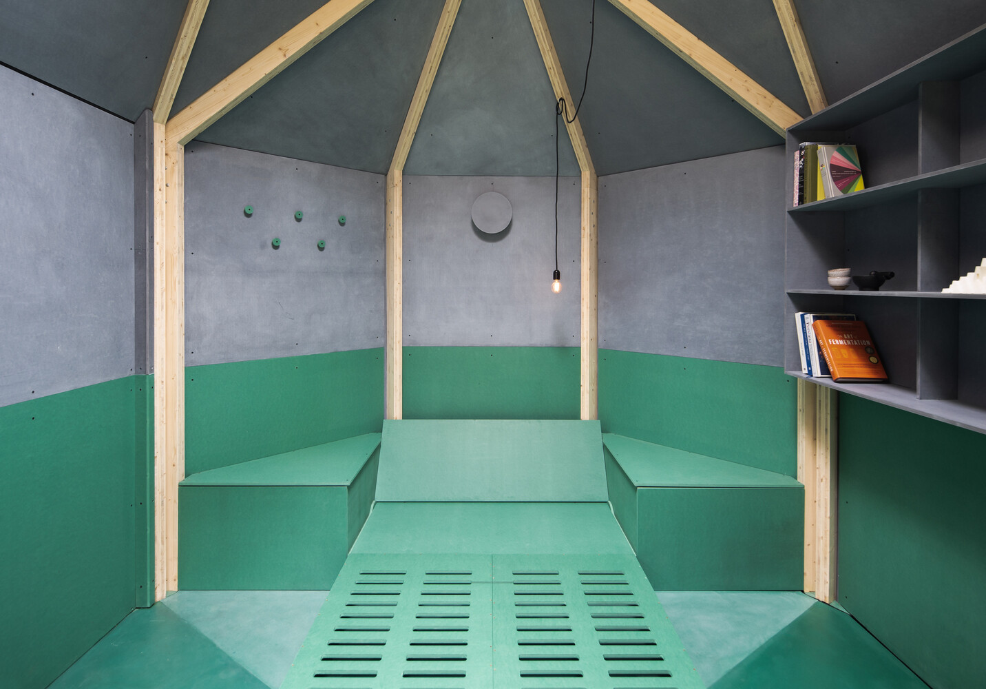 Inside this backyard studio, exposed timber framing converges at the ceiling, creating a sense of height, while angled walls, a built-in desk, and a fold-out bed make the space flexible for work, rest, or quiet contemplation.