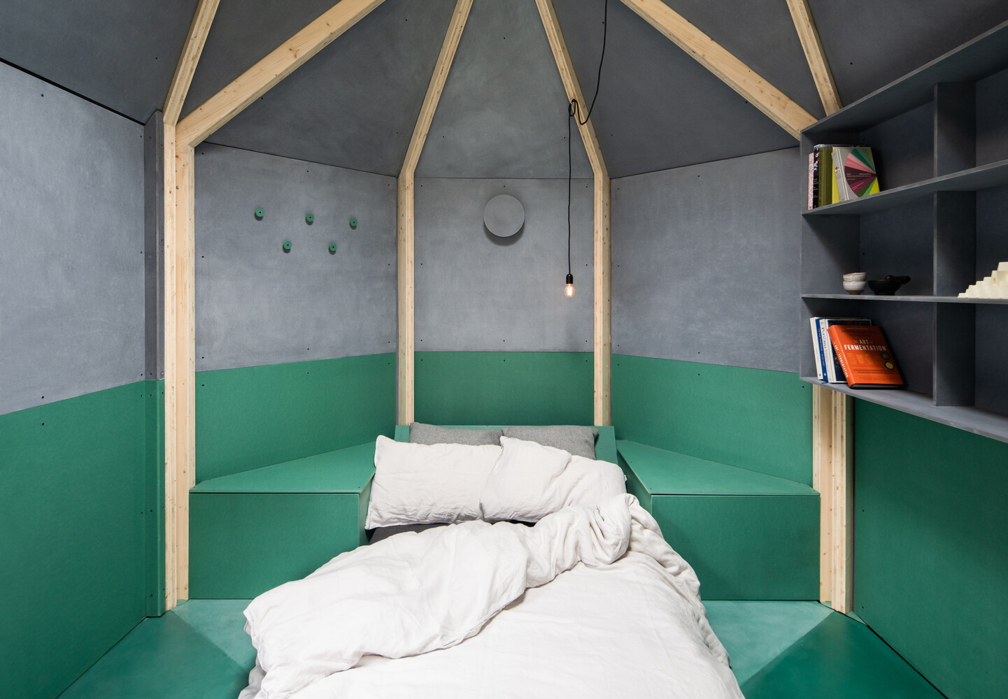 Inside this backyard studio, exposed timber framing converges at the ceiling, creating a sense of height, while angled walls, a built-in desk, and a fold-out bed make the space flexible for work, rest, or quiet contemplation.