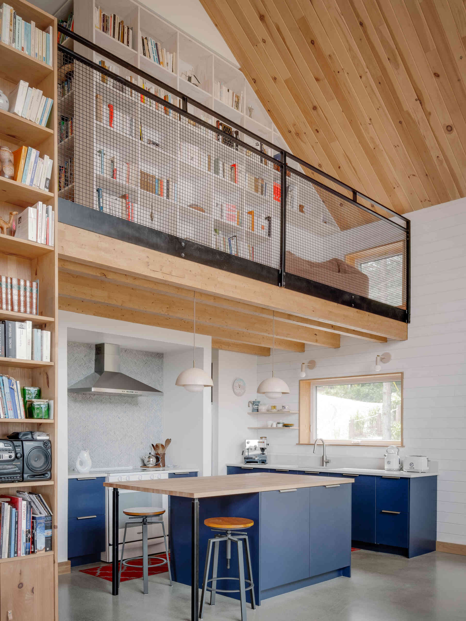 La cocina se ubica ordenadamente en la columna central de la casa, manteniendo todo junto y al mismo tiempo permanece visualmente brillante dentro del espacio de doble altura. Los gabinetes de color azul oscuro marcan el tono de la habitación, contrastando con las paredes blancas y el techo de madera clara, mientras que una simple isla con tapa de madera aporta calidez a la pieza central.