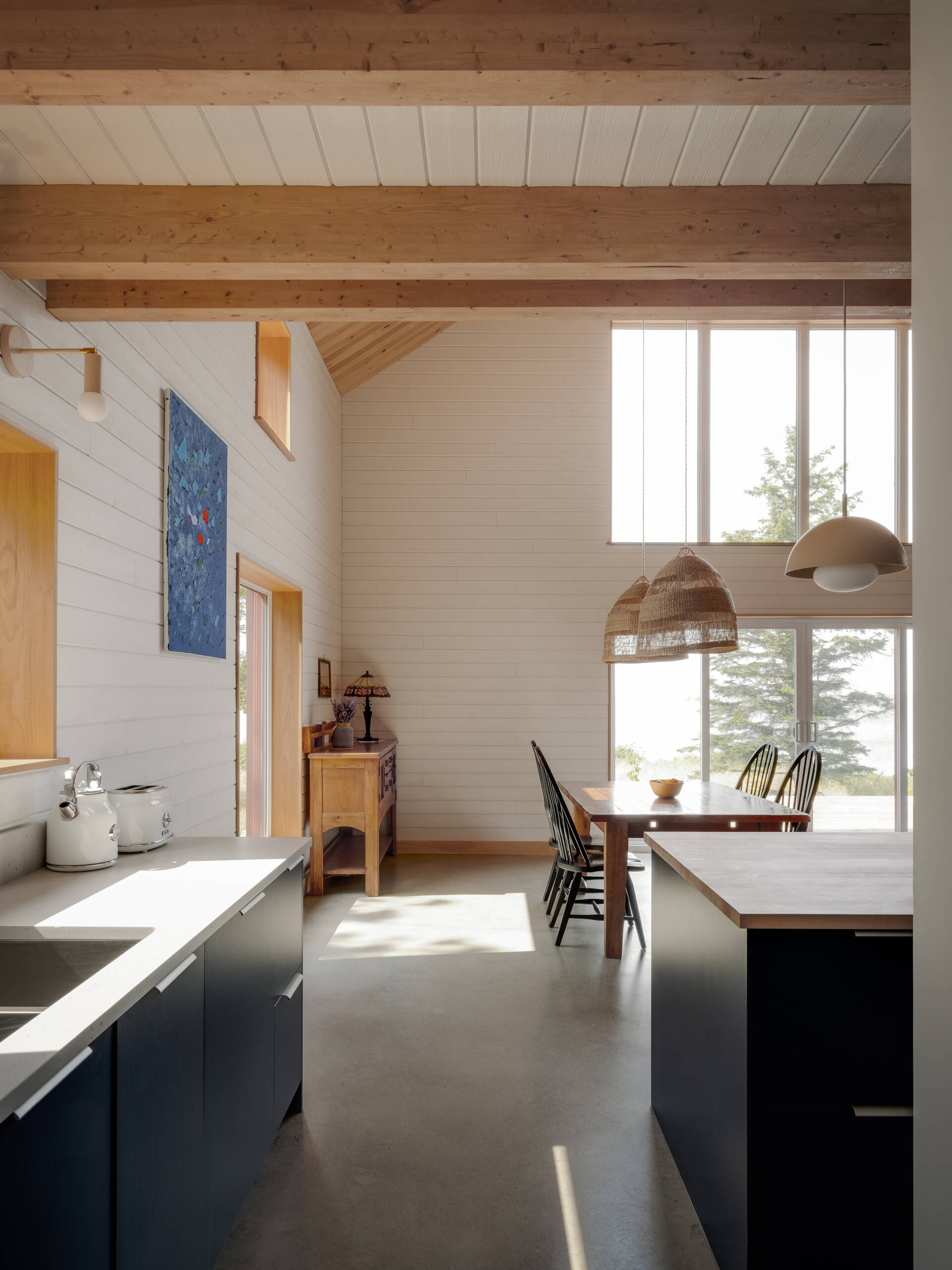 La cocina se ubica ordenadamente en la columna central de la casa, manteniendo todo junto y al mismo tiempo permanece visualmente brillante dentro del espacio de doble altura. Los gabinetes de color azul oscuro marcan el tono de la habitación, contrastando con las paredes blancas y el techo de madera clara, mientras que una simple isla con tapa de madera aporta calidez a la pieza central.