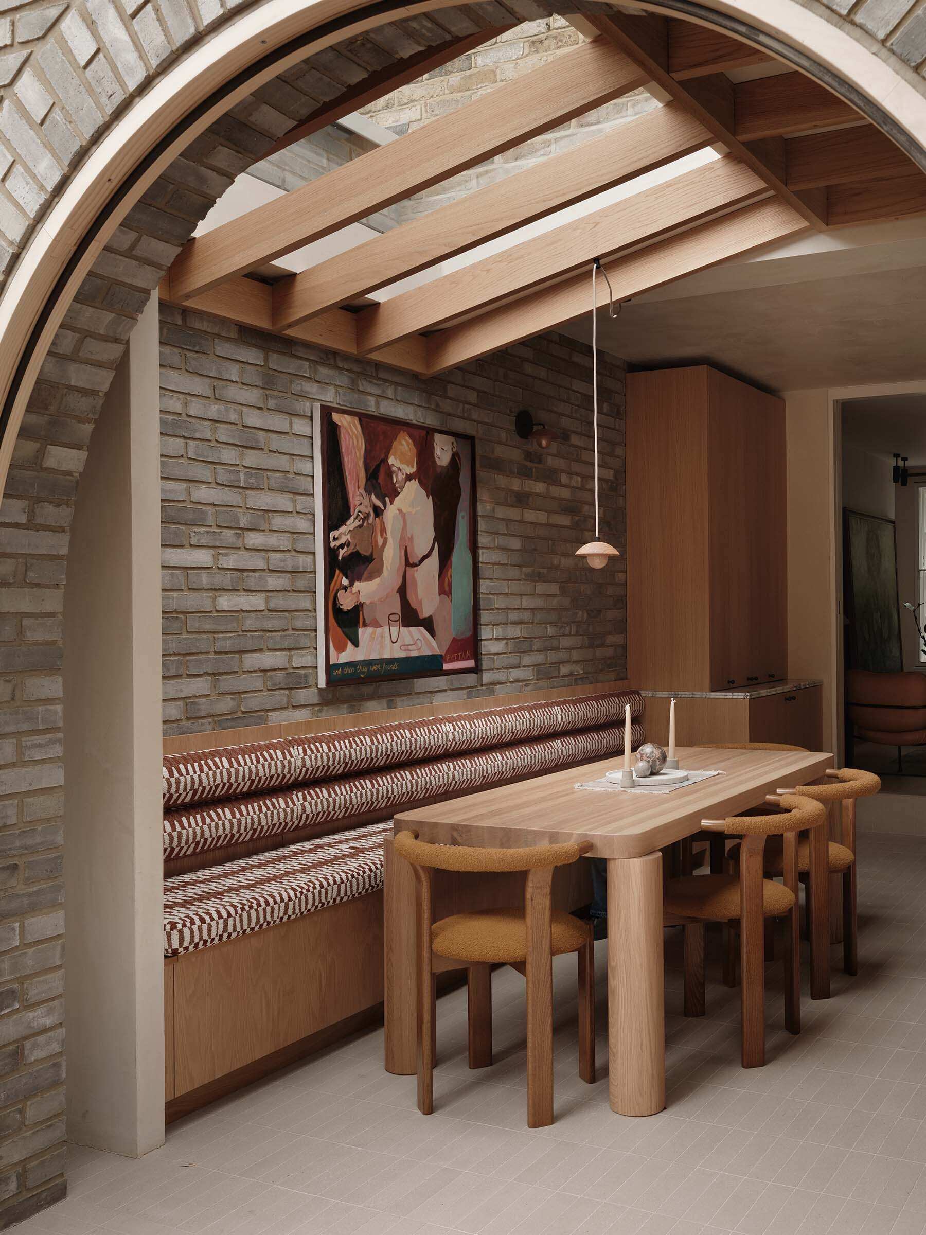 The dining area sits at the center of the open-plan arrangement. It connects directly to both the kitchen and living spaces, allowing it to function as part of a shared environment. The banquette offers seating along the wall, while skylights let natural light to fill the interior.
