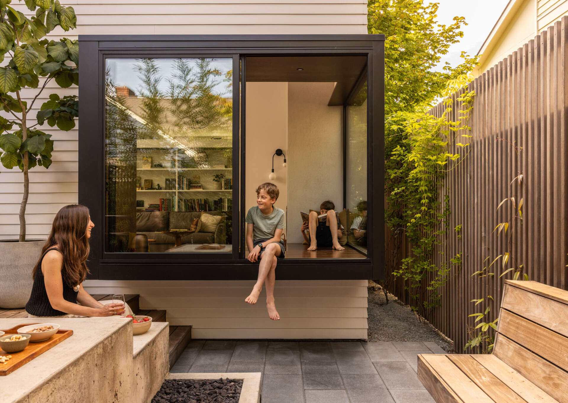 A compact backyard transforms into a layered outdoor retreat with a firepit, built-in seating, and a playful corner window connection.