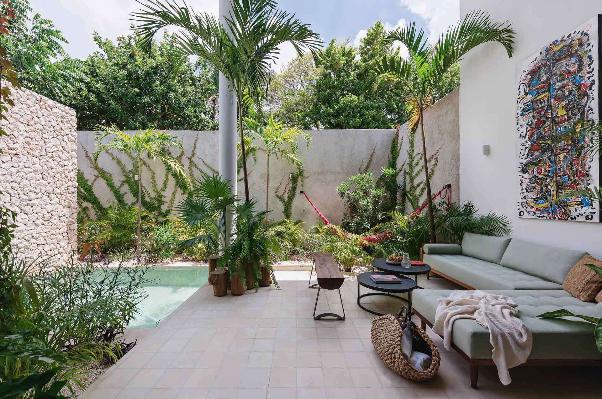 A small courtyard patio filled with tropical plants, colorful artwork, and relaxed seating creates a private outdoor living room inside this modern Mexican home.
