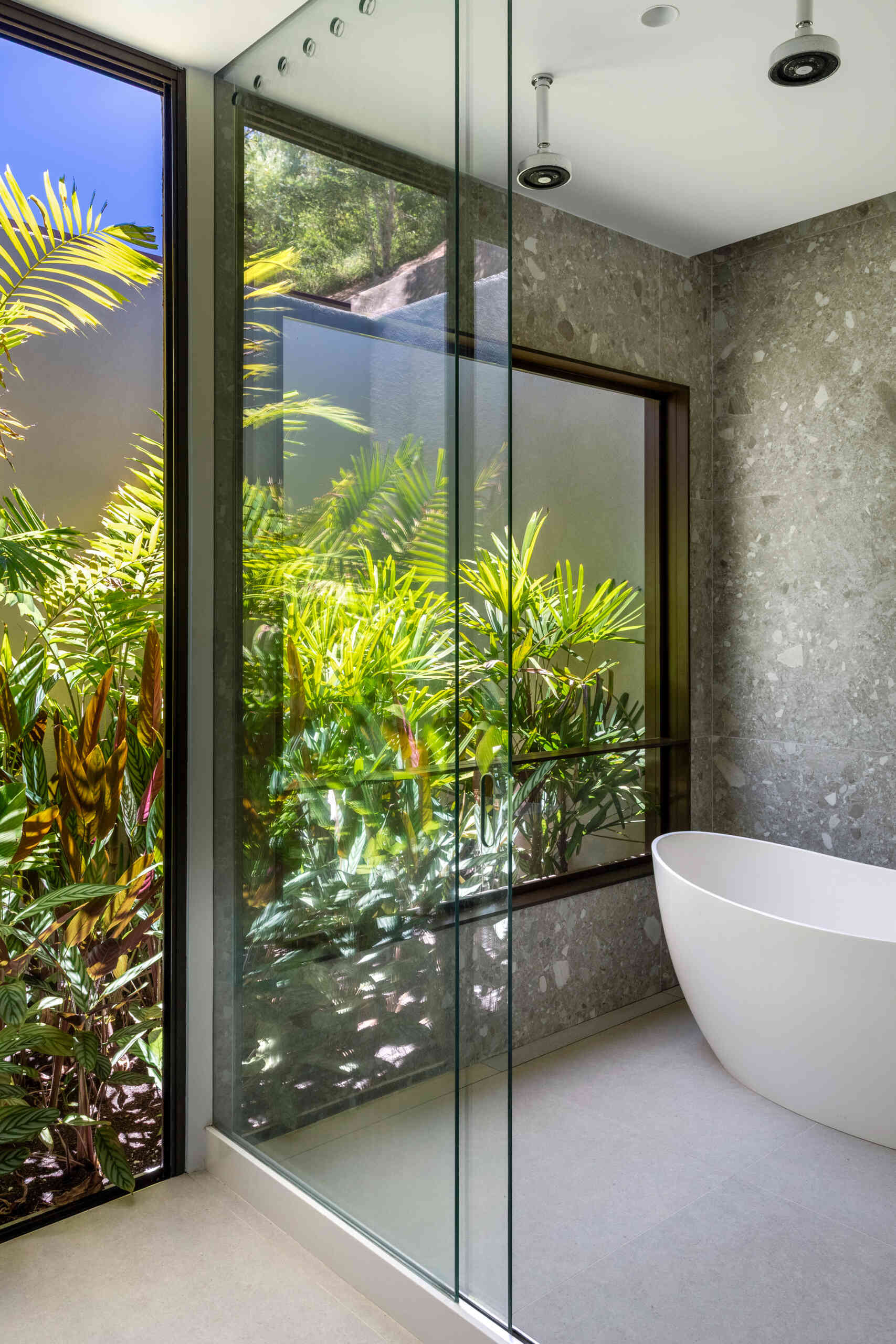 In this bathroom, there’s a feeling of a private garden hideaway. Floor-to-ceiling glass keeps the space open, letting sunlight filter through greenery and cast soft, leafy reflections. Gray stone walls add subtle texture, while a white freestanding tub sits within the wet area.