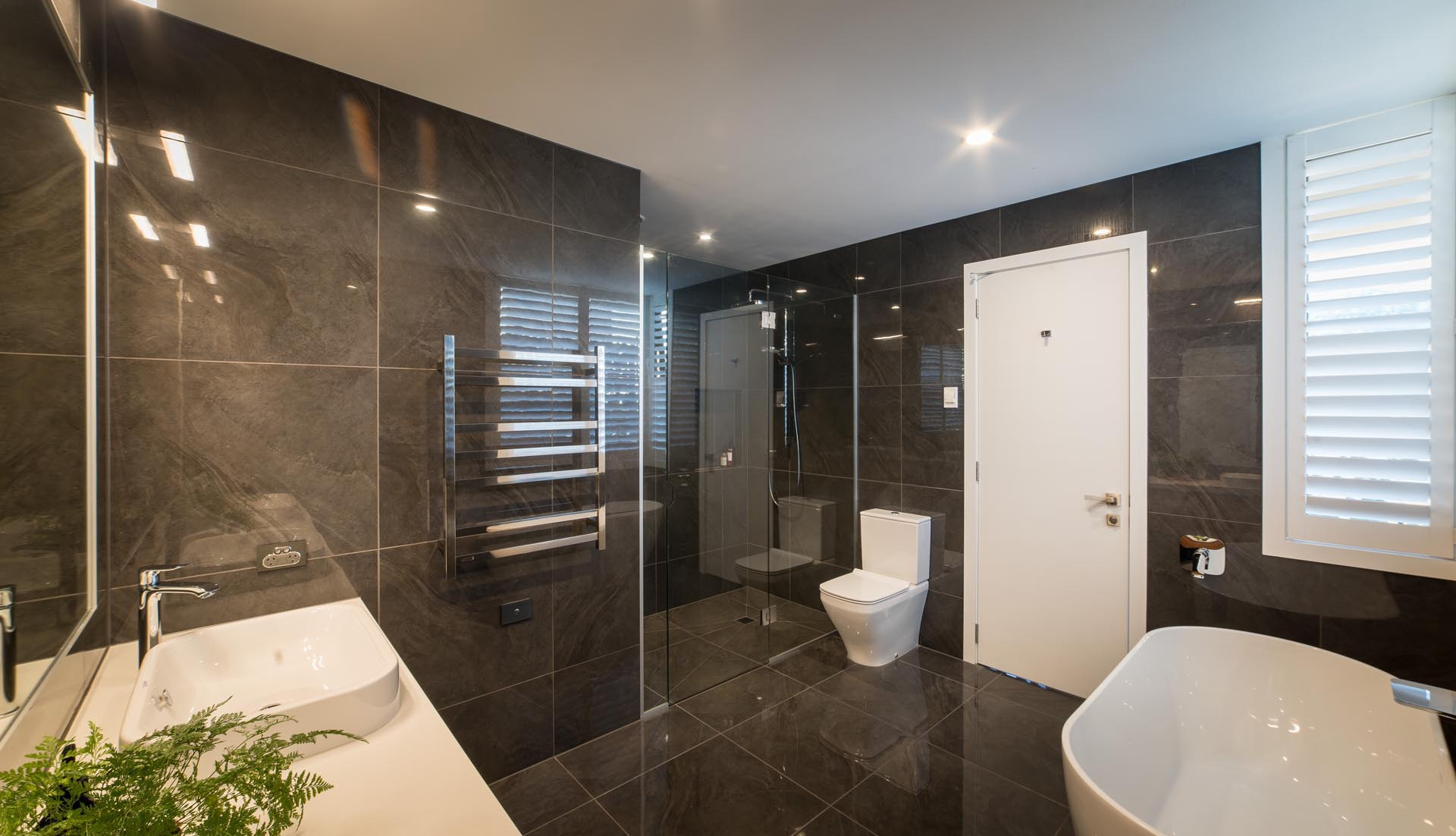 Este baño principal presenta una paleta de colores oscuros con azulejos de gran formato que cubren las paredes y el piso. La bañera y la encimera independientes de color blanco contrastan con las superficies oscuras para crear un punto focal llamativo.