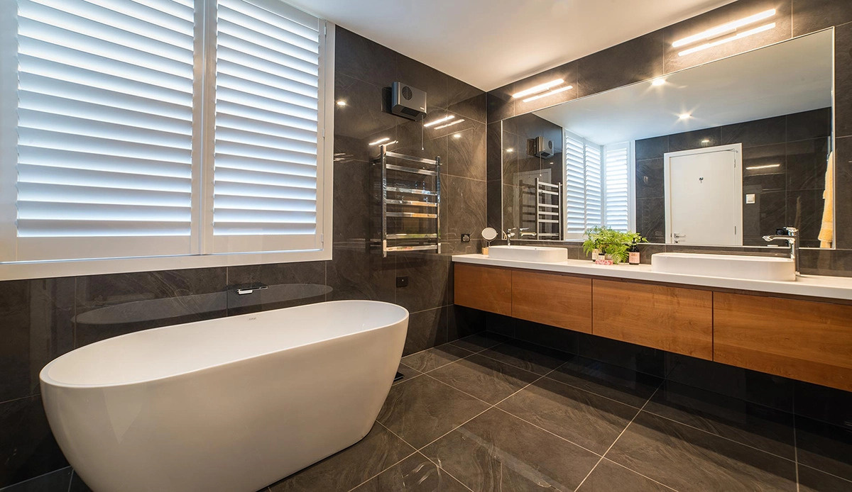 Este baño principal presenta una paleta de colores oscuros con azulejos de gran formato que cubren las paredes y el piso. La bañera y la encimera independientes de color blanco contrastan con las superficies oscuras para crear un punto focal llamativo.