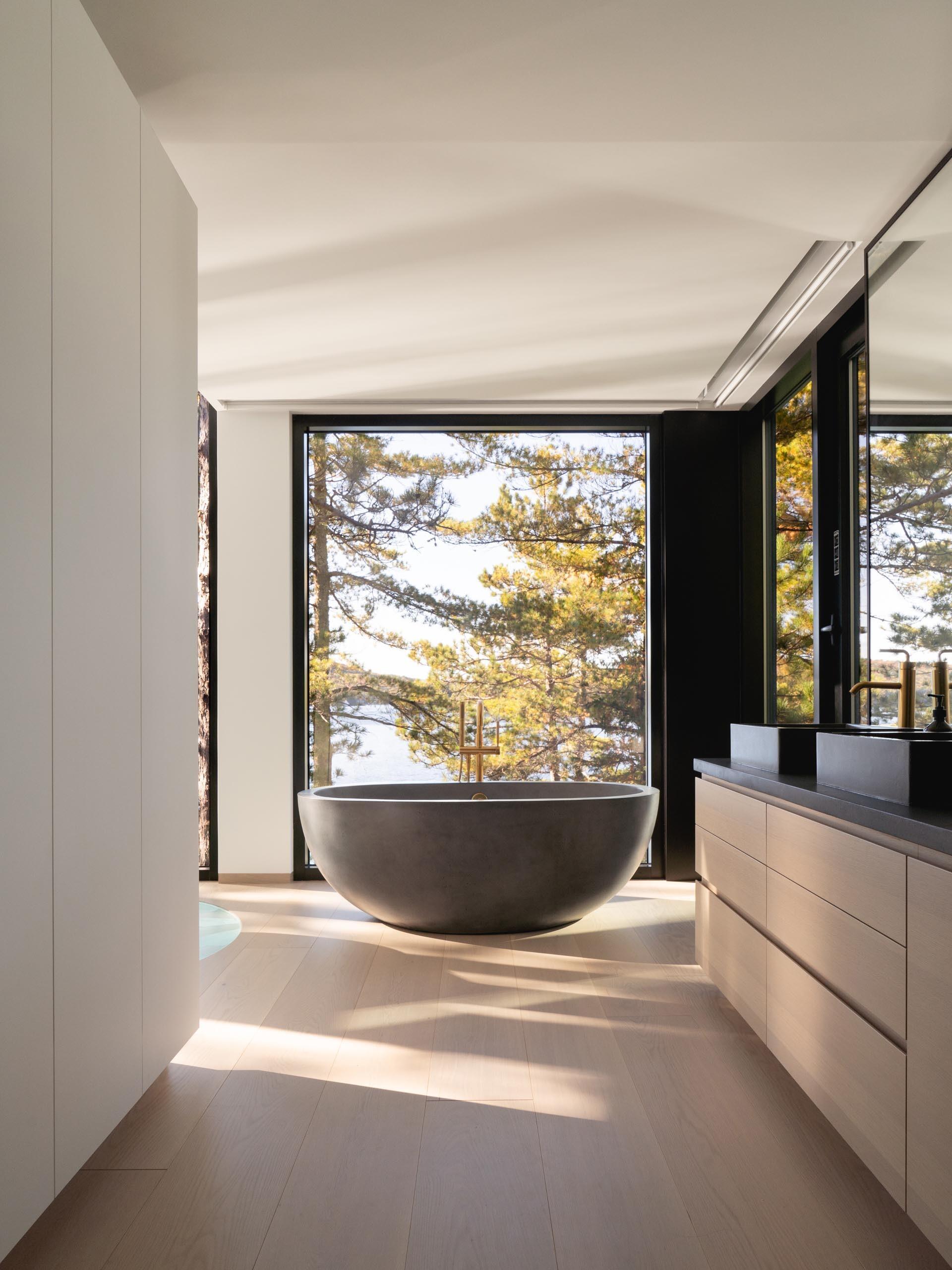 The primary bedroom features floor-to-ceiling windows, a suspended black fireplace, and a freestanding bathtub that frames sweeping views of the Quebec lake and surrounding pines.