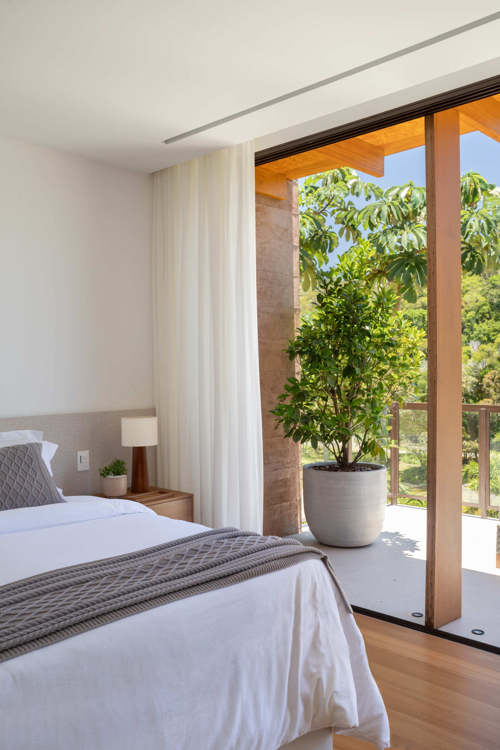 This bedroom is calm and inviting, with warm wood accents and soft textures. Sheer curtains filter sunlight, and wide openings to a private outdoor space.