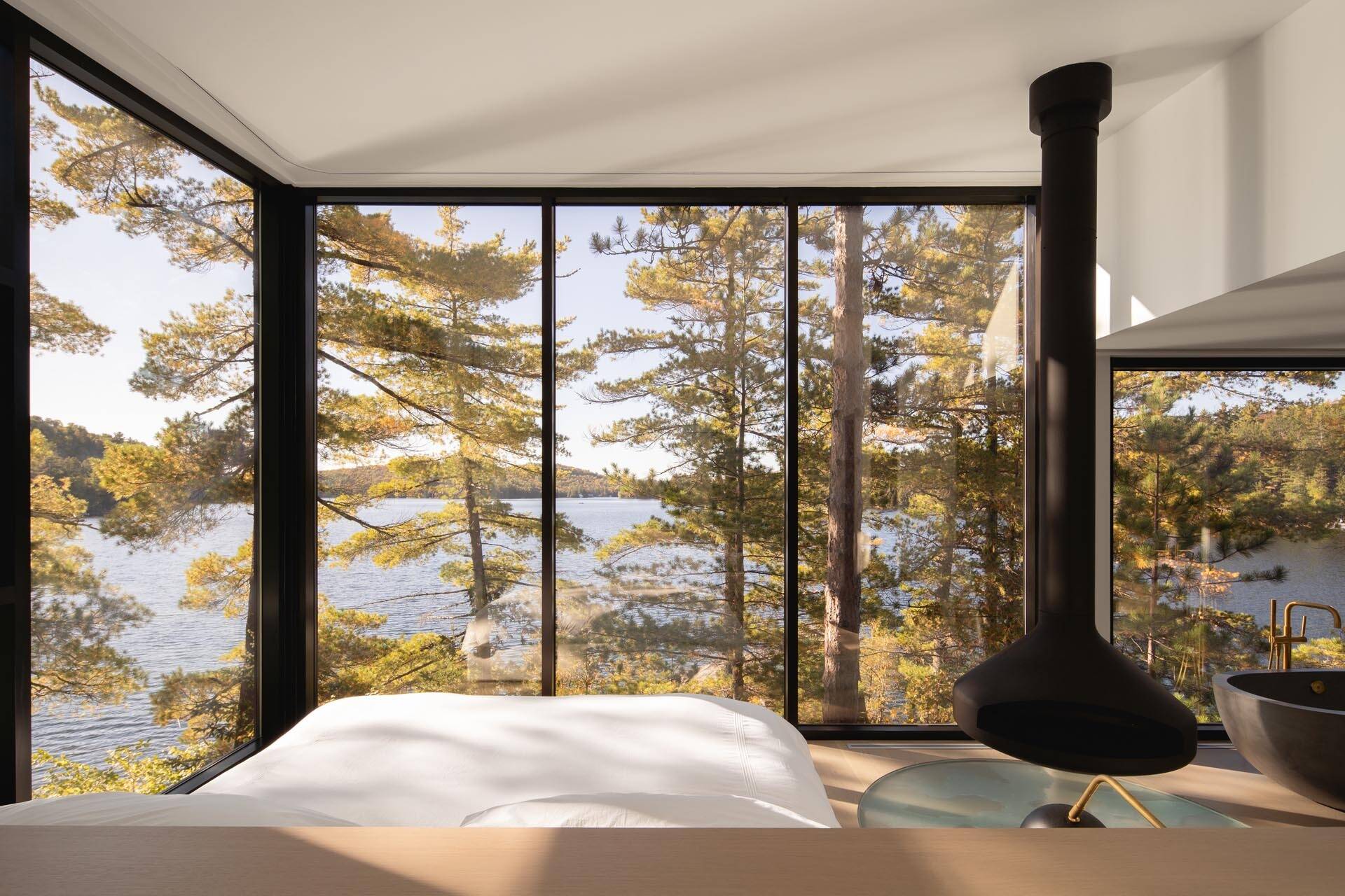 The primary bedroom features floor-to-ceiling windows, a suspended black fireplace, and a freestanding bathtub that frames sweeping views of the Quebec lake and surrounding pines.