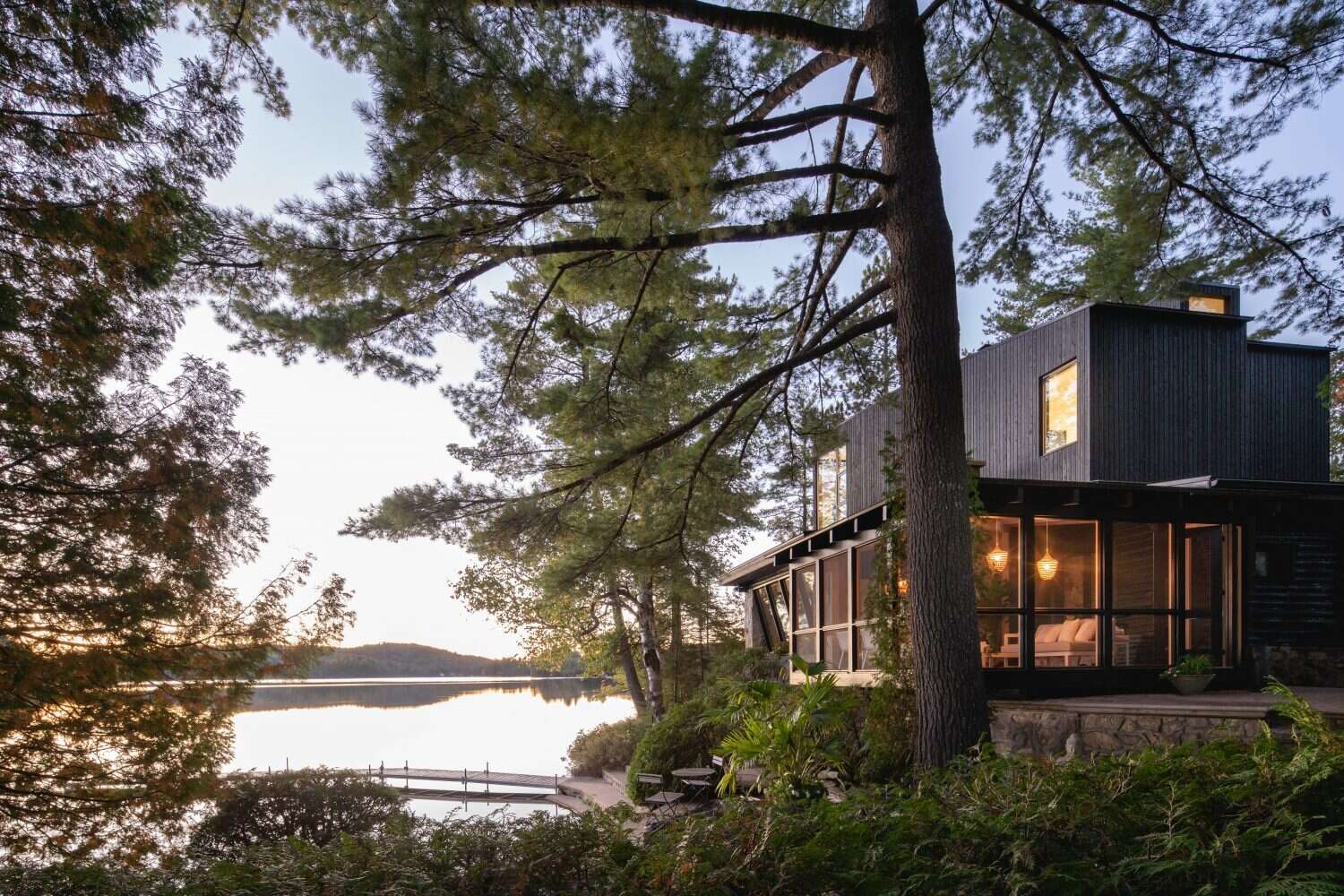 A log cabin on a rocky point in Quebec was given a contemporary second life by Paul Bernier Architecte. The remodel adds a second level and a screened porch while preserving the rustic character of the original chalet. A dark exterior and stone foundation help the cabin blend into the surrounding landscape and lakeshore.