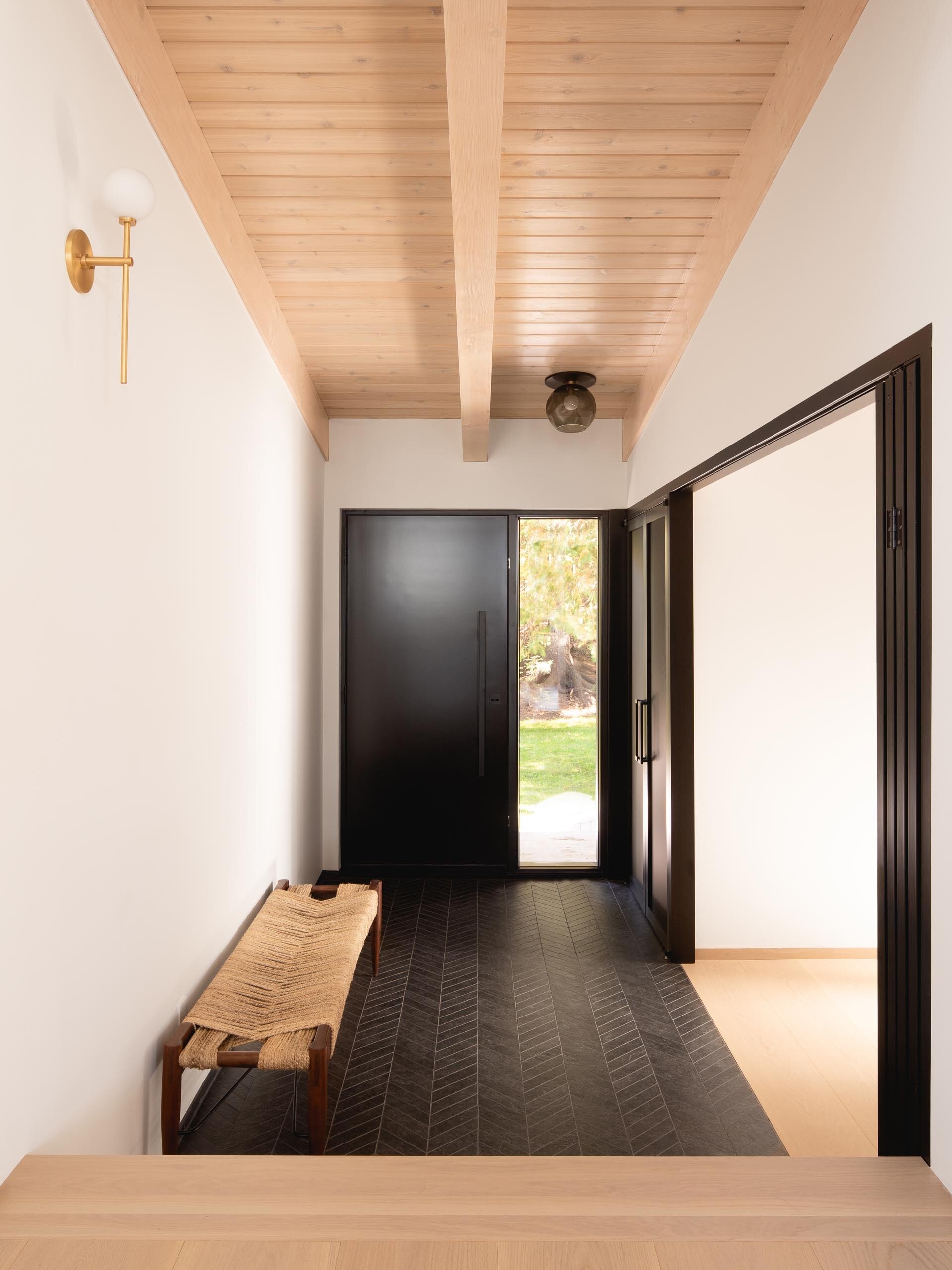 A bright entryway welcomes guests into this renovated Quebec log cabin, where a light wood ceiling contrasts with a bold black chevron tile floor that hints at the home’s modern transformation.