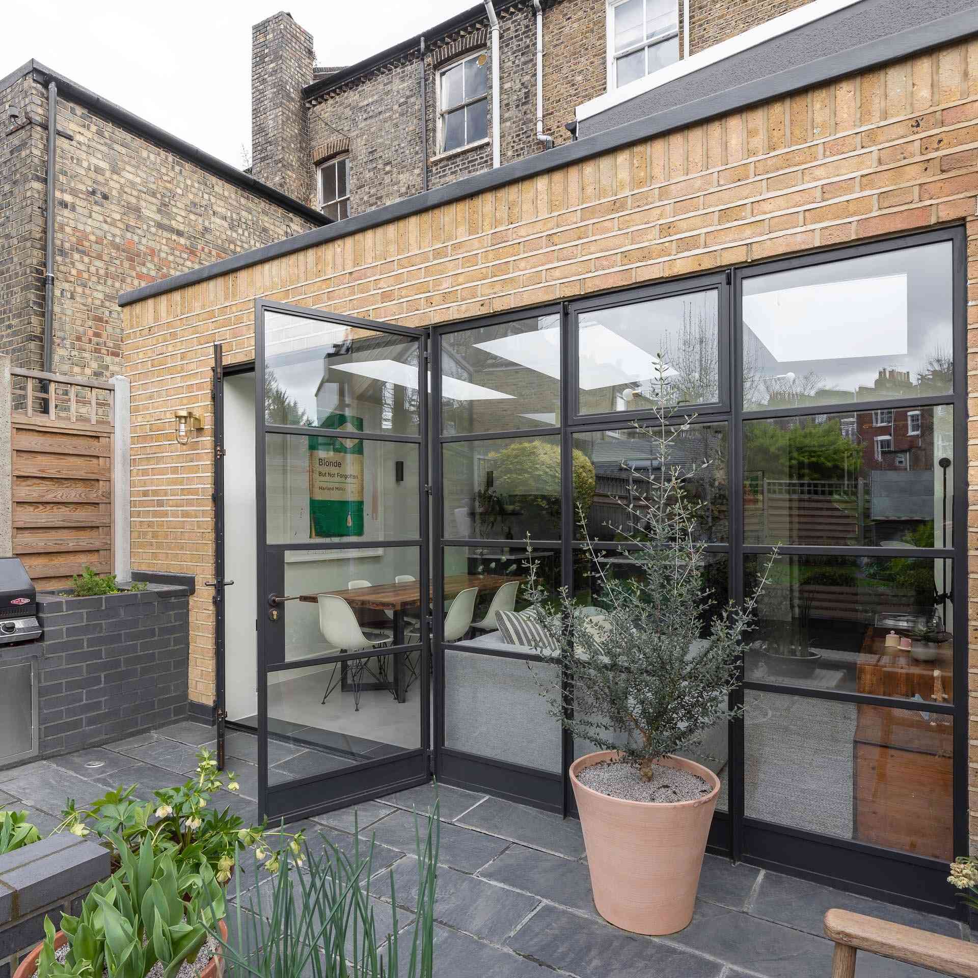 A black framed glass wall opens the extension to a patio with seating and an outdoor kitchen with a BBQ, creating a seamless indoor outdoor connection.