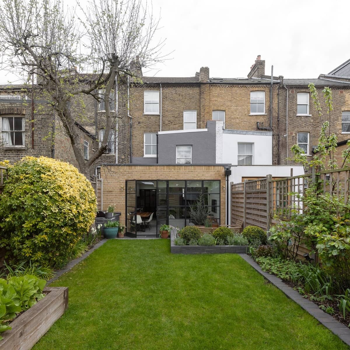 A black framed glass wall opens the extension to a patio with seating and an outdoor kitchen with a BBQ, creating a seamless indoor outdoor connection.