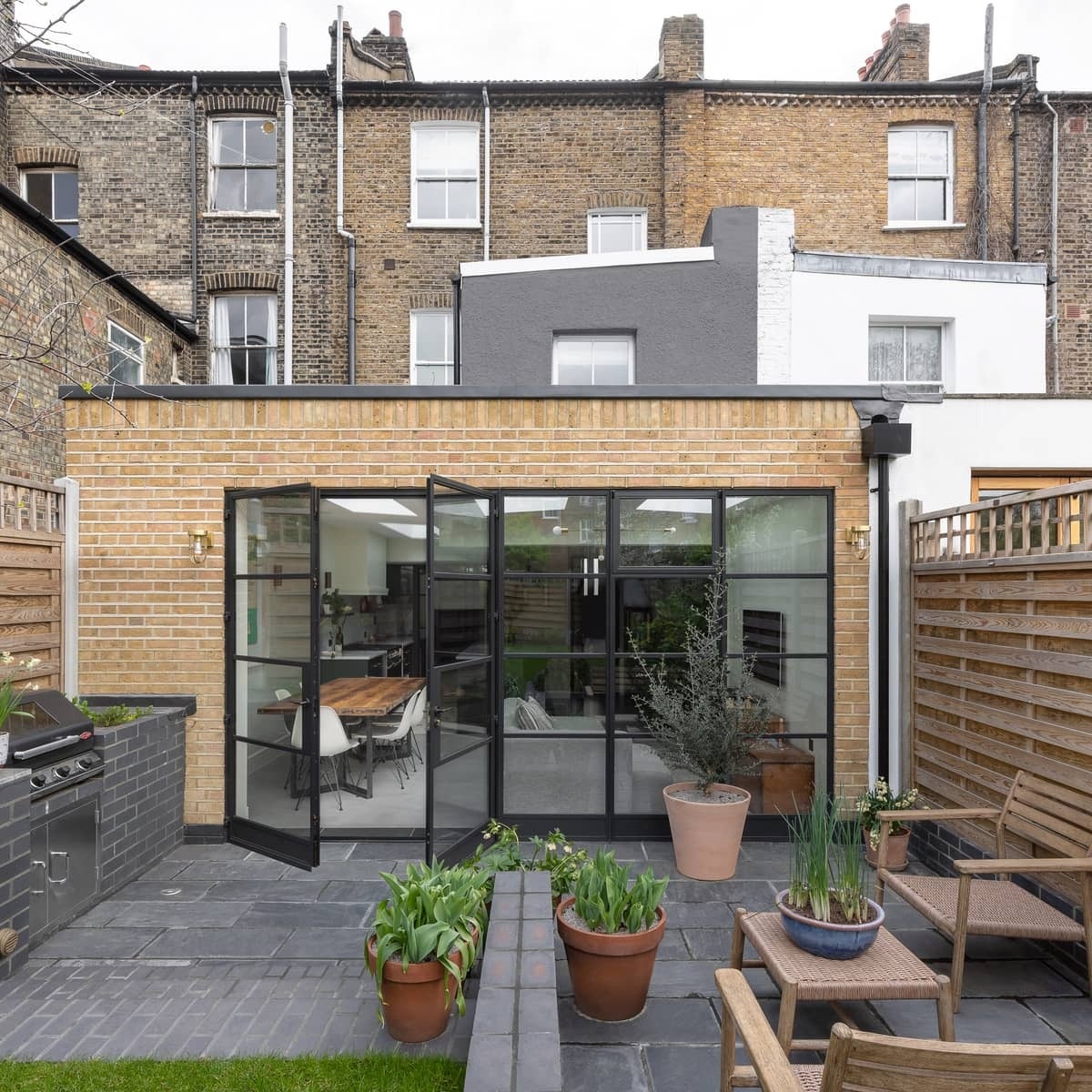 A black framed glass wall opens the extension to a patio with seating and an outdoor kitchen with a BBQ, creating a seamless indoor outdoor connection.