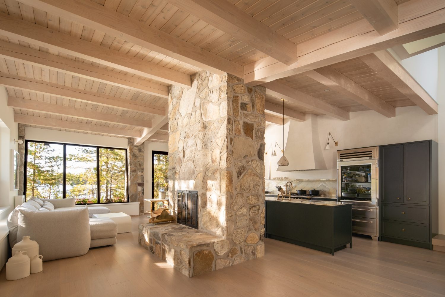 A U-shaped sofa in the living room faces lake views while a stone fireplace anchors the space and subtly separates it from the kitchen beyond.