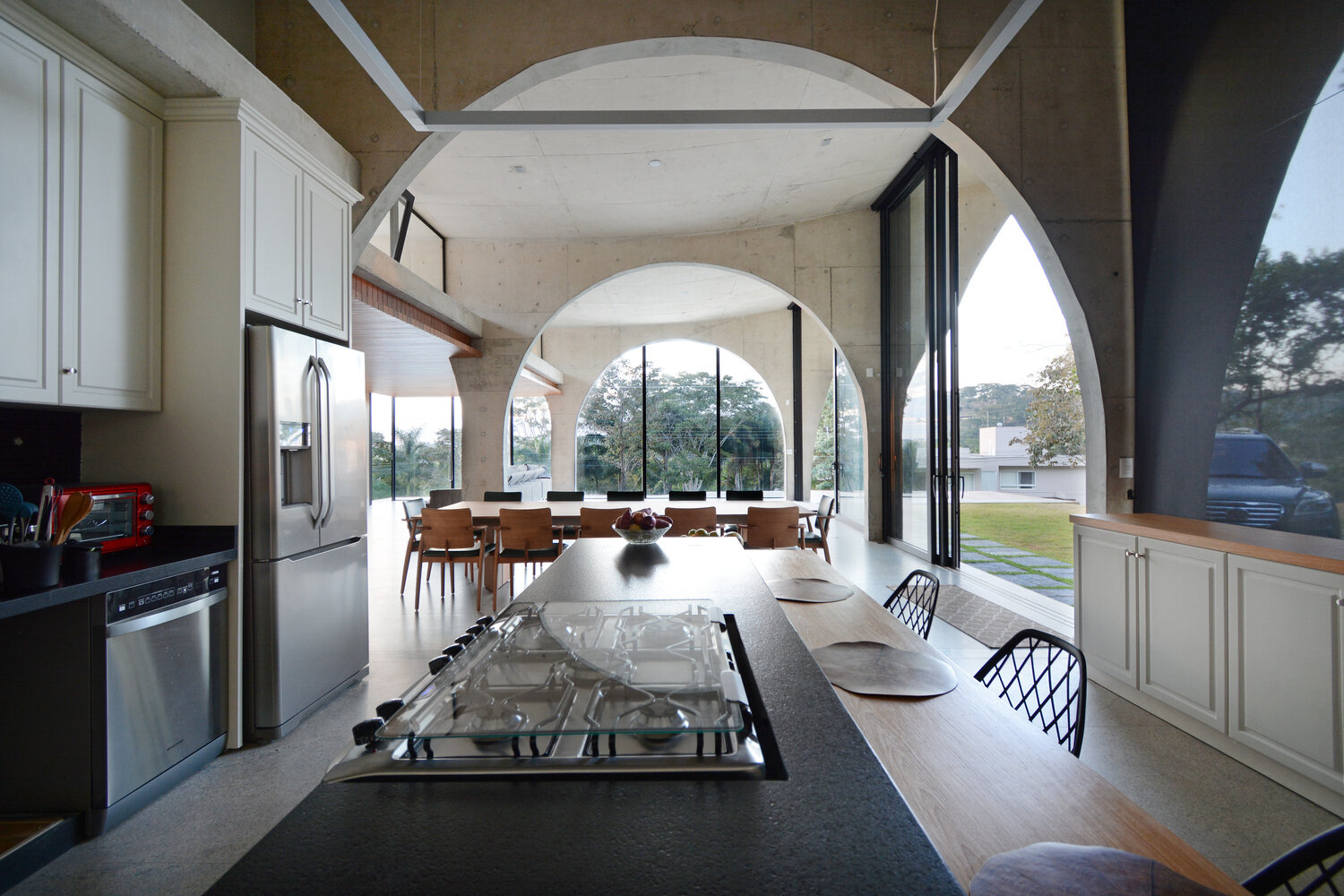 The living room, dining area, and kitchen share a dramatic double height space framed by concrete arches and large sliding glass doors that connect the interior to the gardens.