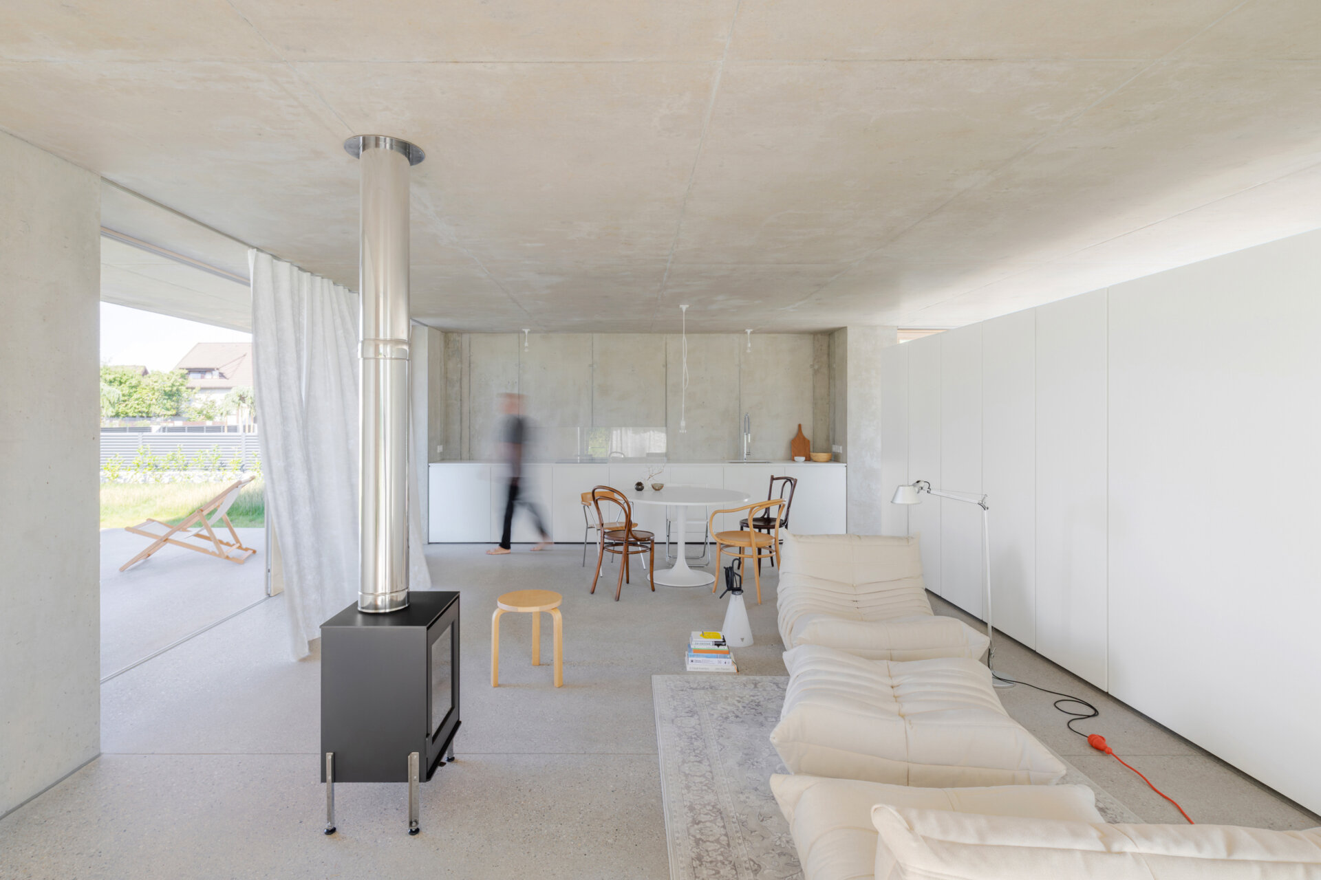 An open plan living, dining and kitchen layout with polished concrete floors, a cast iron fireplace, and a double sided storage wall separating the bedroom and living zones.