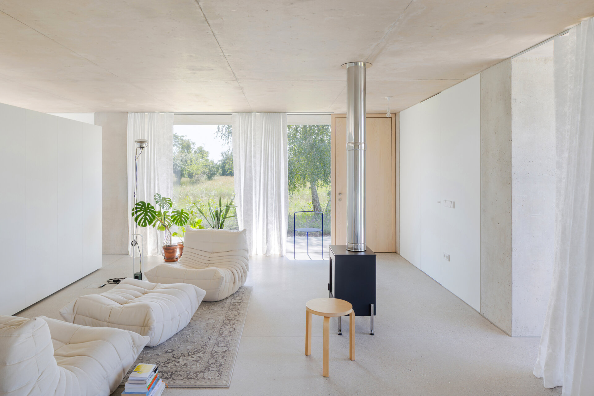An open plan living, dining and kitchen layout with polished concrete floors, a cast iron fireplace, and a double sided storage wall separating the bedroom and living zones.