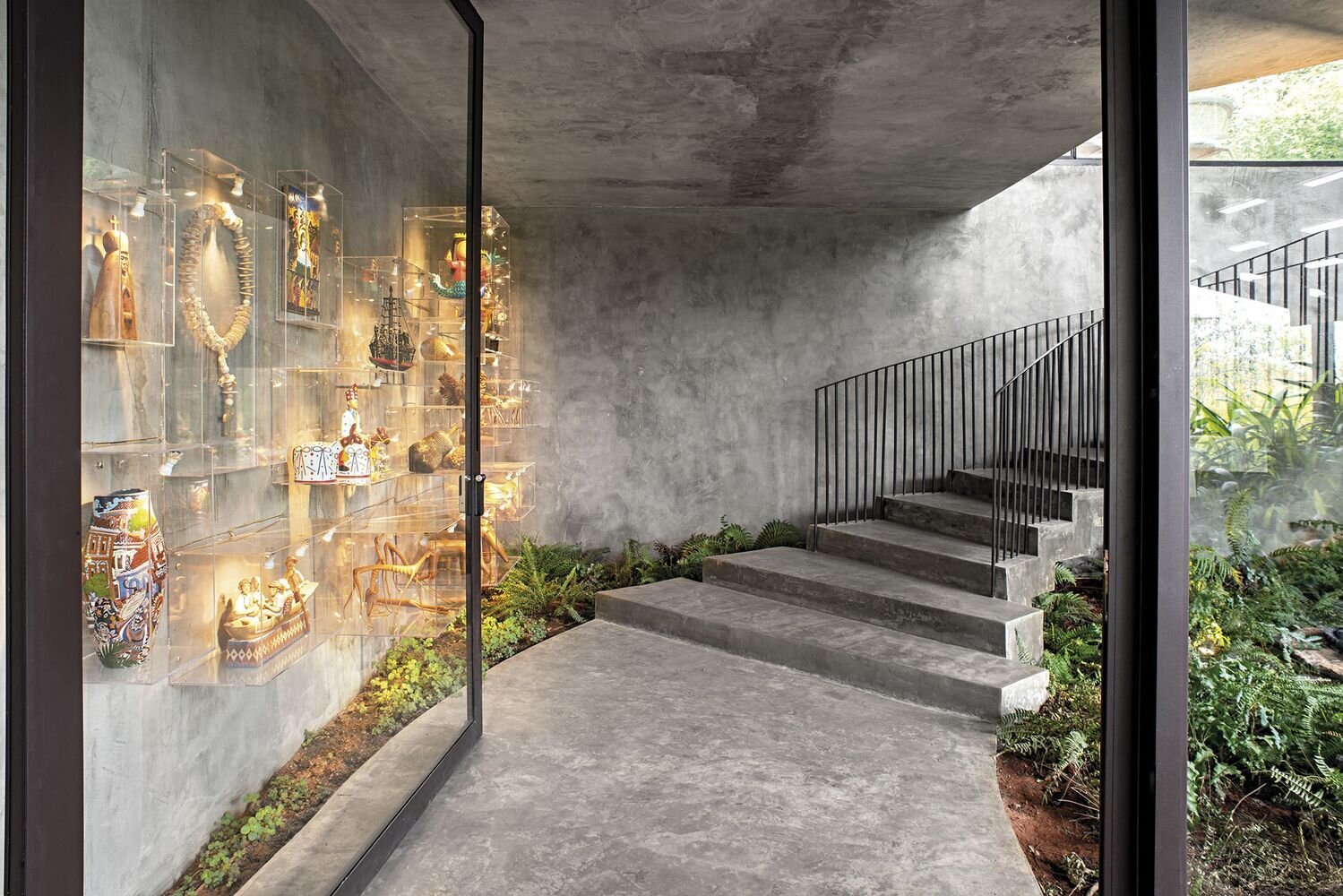 Arrival at the home is marked by a sequence of strong visual elements. An oversized black-framed glass door sets the tone, paired with stairs finished with black railings.

Along the way, a wall with clear floating boxes display sculptural pieces, while surrounding gardens soften the experience. Together, these details create a layered and striking entrance that blends architecture with curated moments.