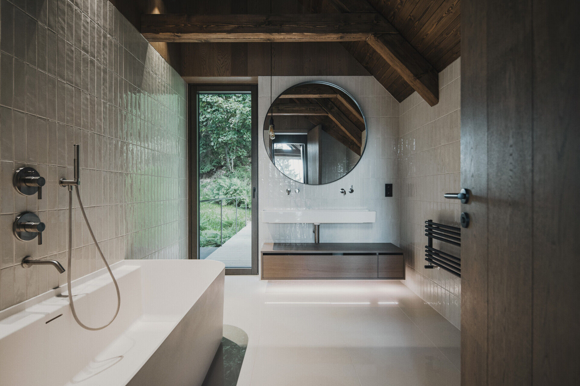 The main bathrooms in the home have glossy off-white tiles wrapping the walls, while one has a freestanding bathtub, and another has simple glass shower. Dark wood accents and exposed beams warm up the vaulted ceiling.