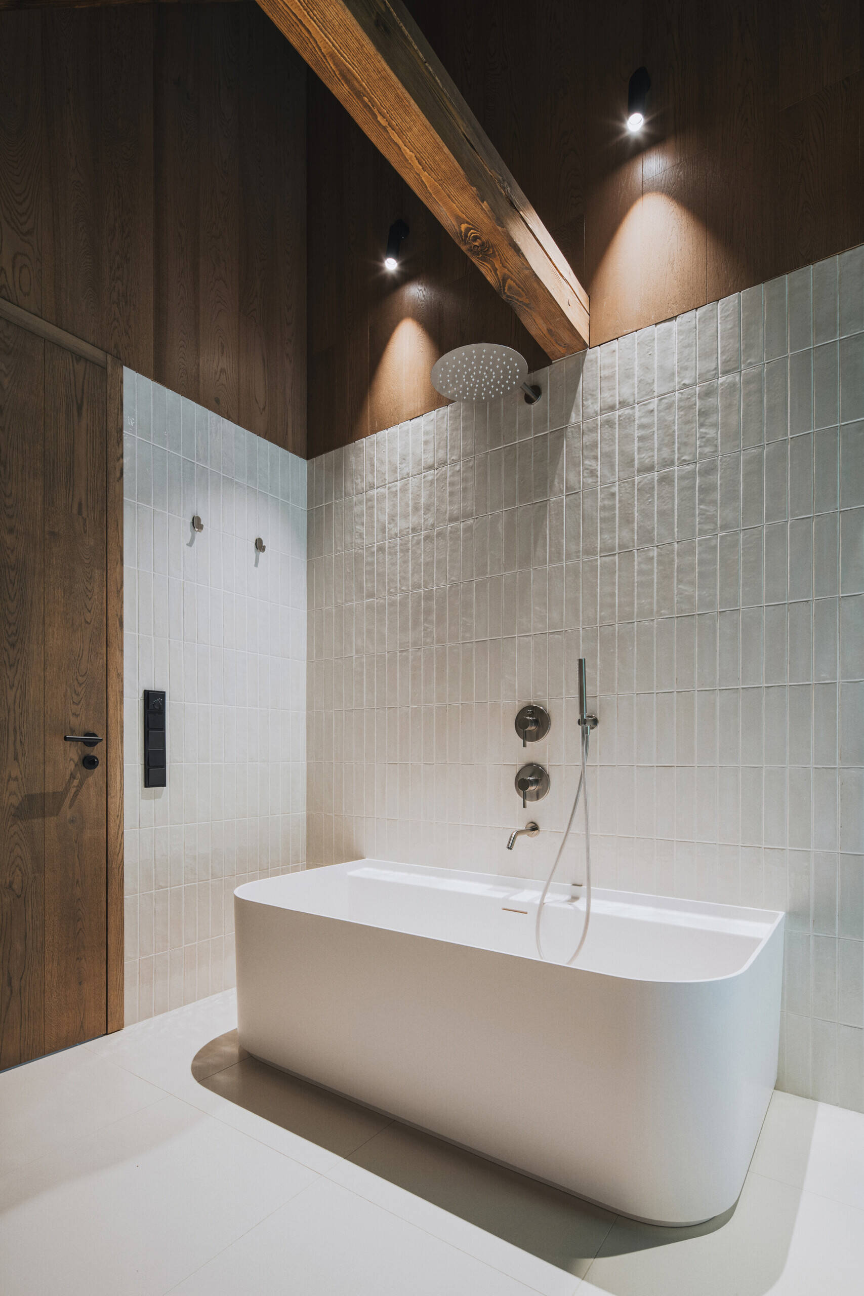 The main bathrooms in the home have glossy off-white tiles wrapping the walls, while one has a freestanding bathtub, and another has simple glass shower. Dark wood accents and exposed beams warm up the vaulted ceiling.