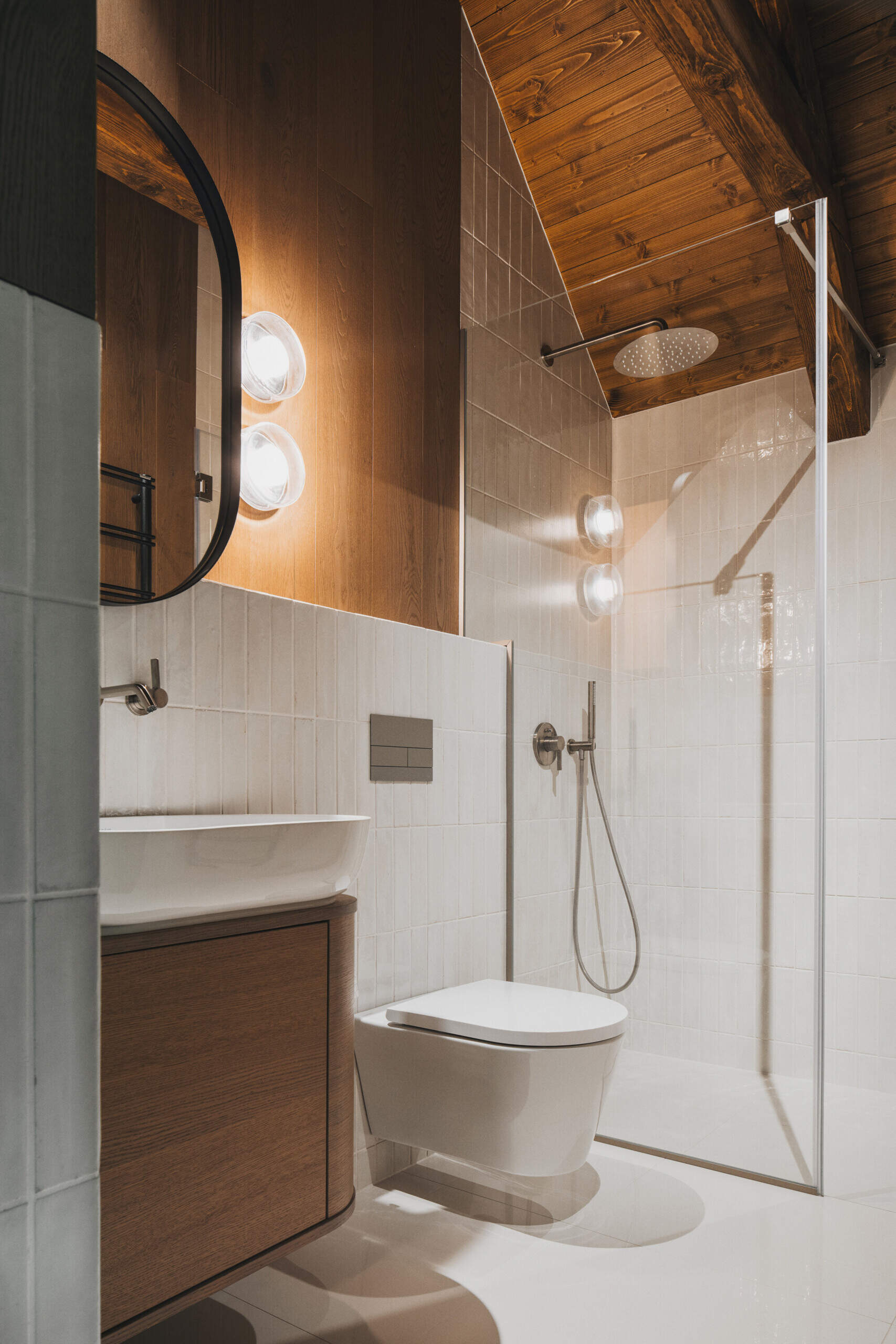The main bathrooms in the home have glossy off-white tiles wrapping the walls, while one has a freestanding bathtub, and another has simple glass shower. Dark wood accents and exposed beams warm up the vaulted ceiling.