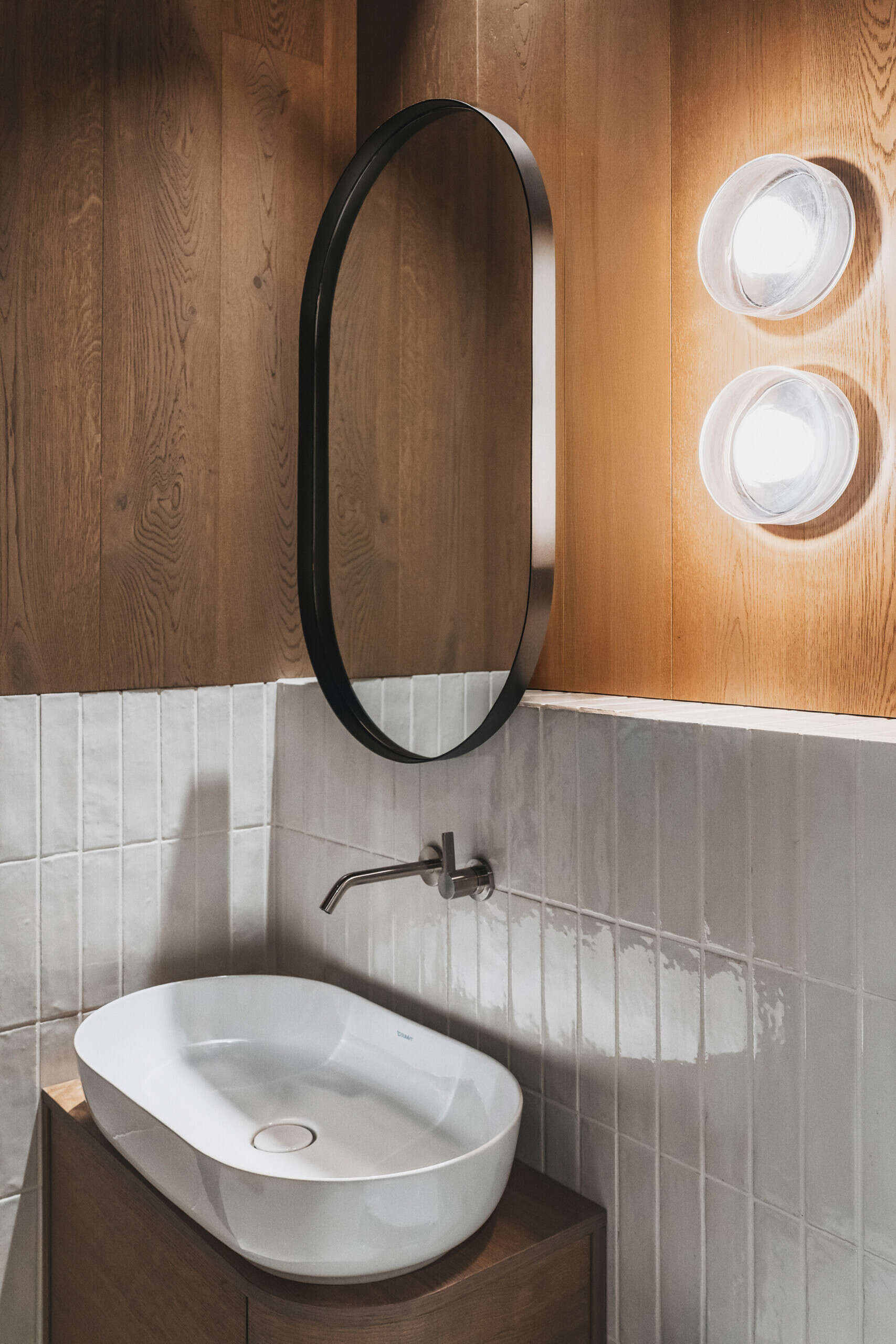 The main bathrooms in the home have glossy off-white tiles wrapping the walls, while one has a freestanding bathtub, and another has simple glass shower. Dark wood accents and exposed beams warm up the vaulted ceiling.