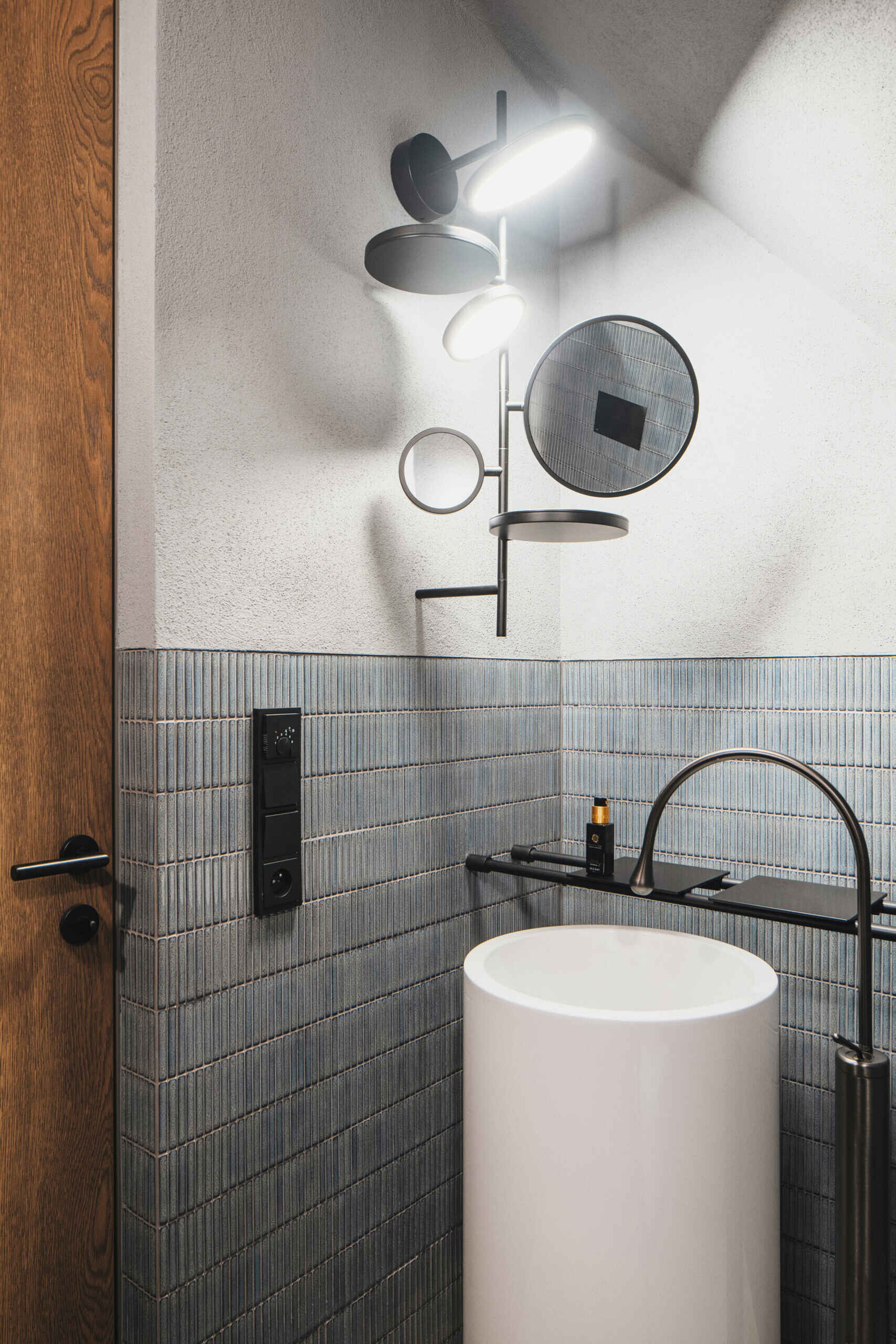 A compact powder room packs in a lot of personality, pairing cool blue-gray tile with a softly textured plaster wall above. The sculptural wall sconce feels like modern jewelry, casting bright, focused light that makes quick handwashing and touch-ups easy.