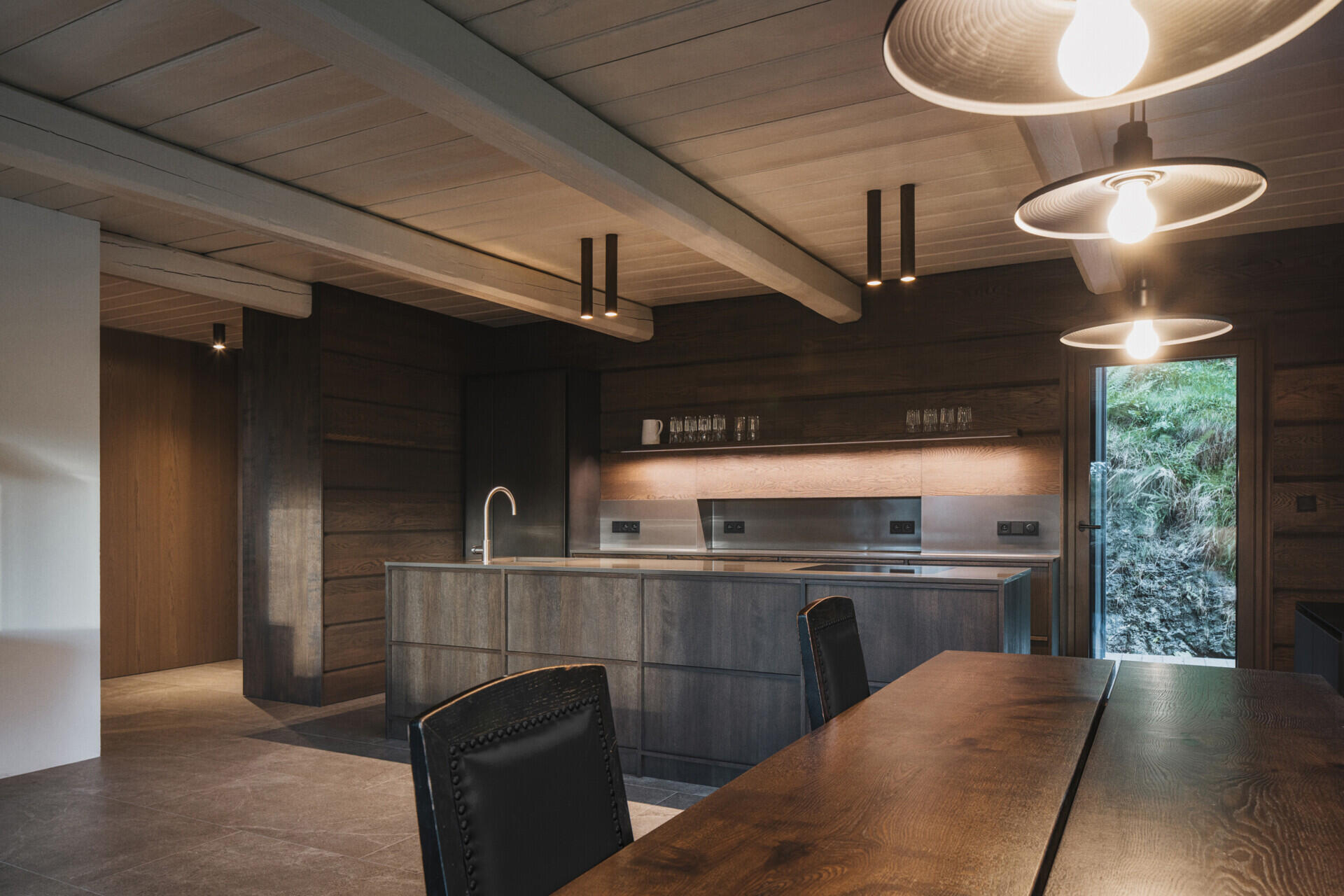 At the heart of this mountain cottage, the main living room retains the scale dictated by an original beam ceiling. Built-in seating runs along the windows, and a large table anchors the space for meals and gatherings. Nearby, the kitchen with its dark cabinetry, almost blends into the background.