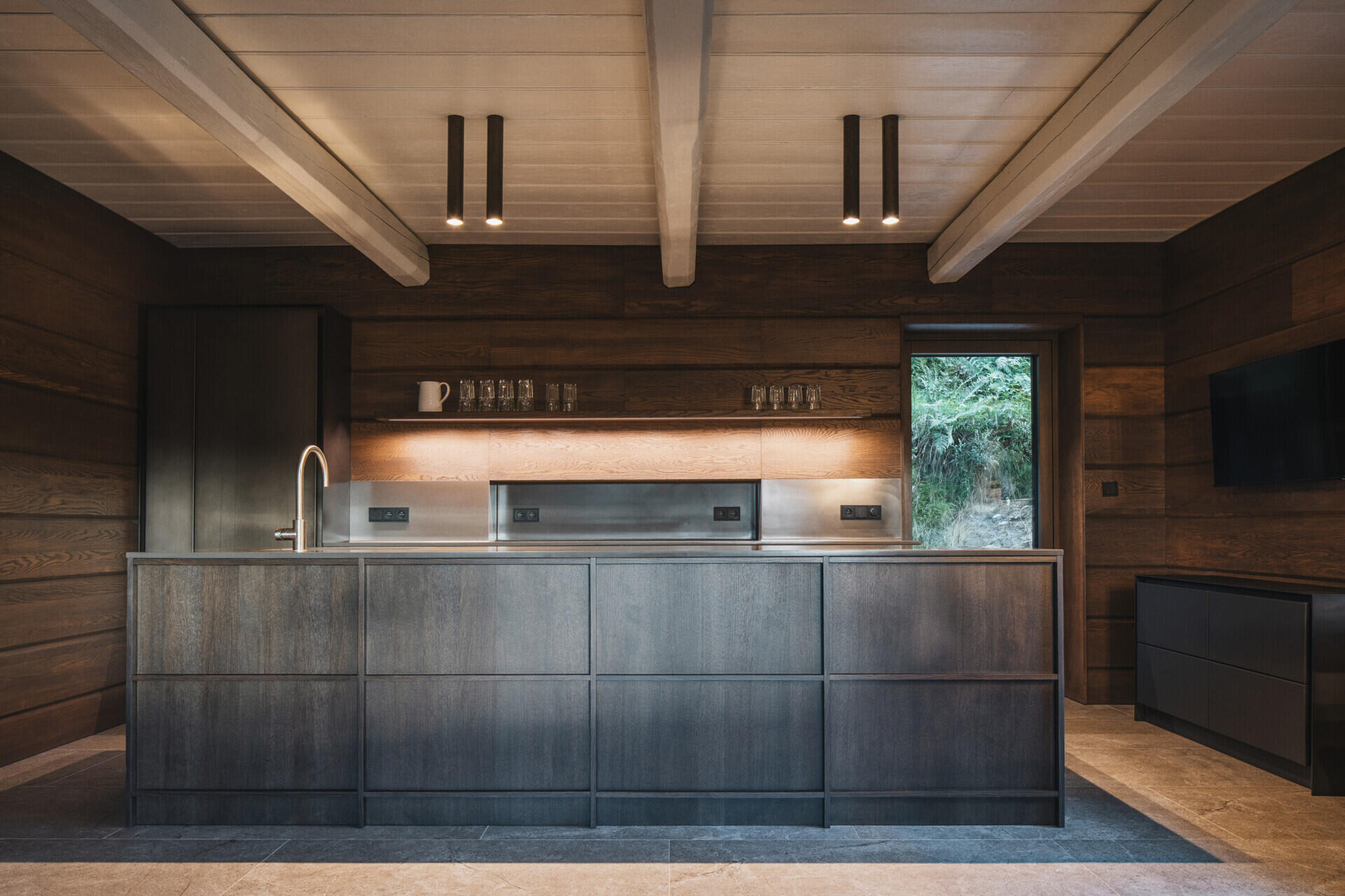 At the heart of this mountain cottage, the main living room retains the scale dictated by an original beam ceiling. Built-in seating runs along the windows, and a large table anchors the space for meals and gatherings. Nearby, the kitchen with its dark cabinetry, almost blends into the background.