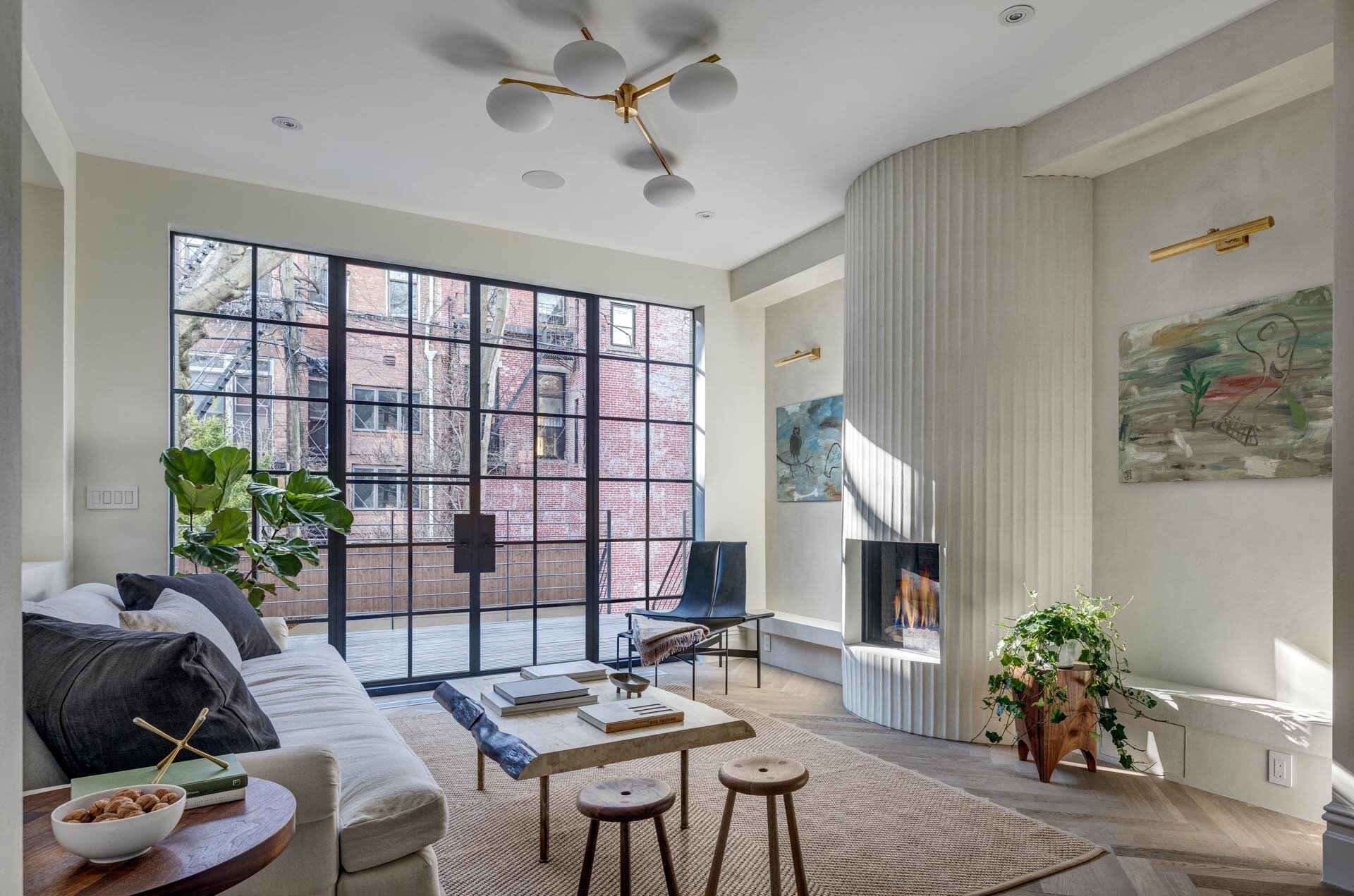 A floor to ceiling fluted plaster fireplace transforms this 1860s Brooklyn living room into a sculptural focal point, balanced by steel doors, built in shelving, and curated art.