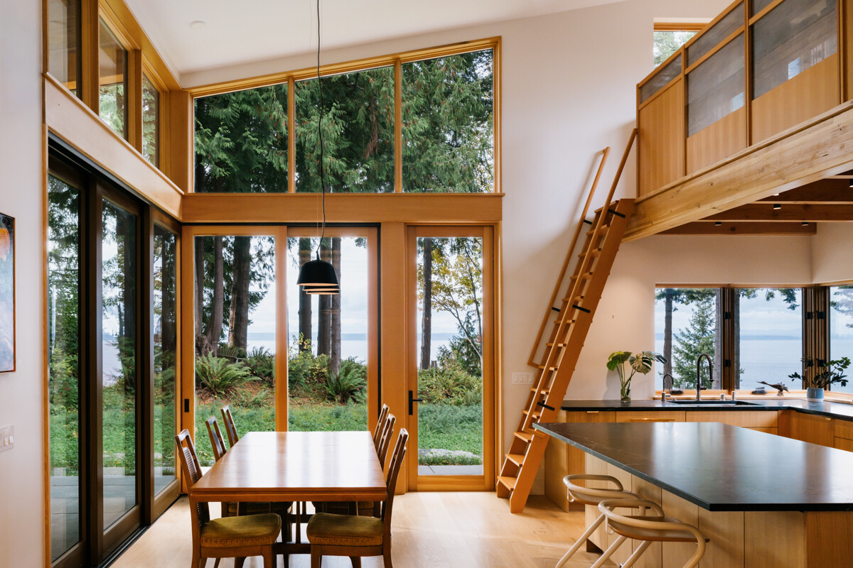 A double-height glass corner dining space that captures sweeping views of trees, water, and sky.