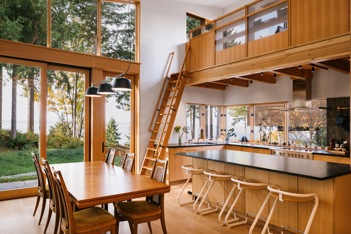 A double-height glass corner dining space that captures sweeping views of trees, water, and sky.