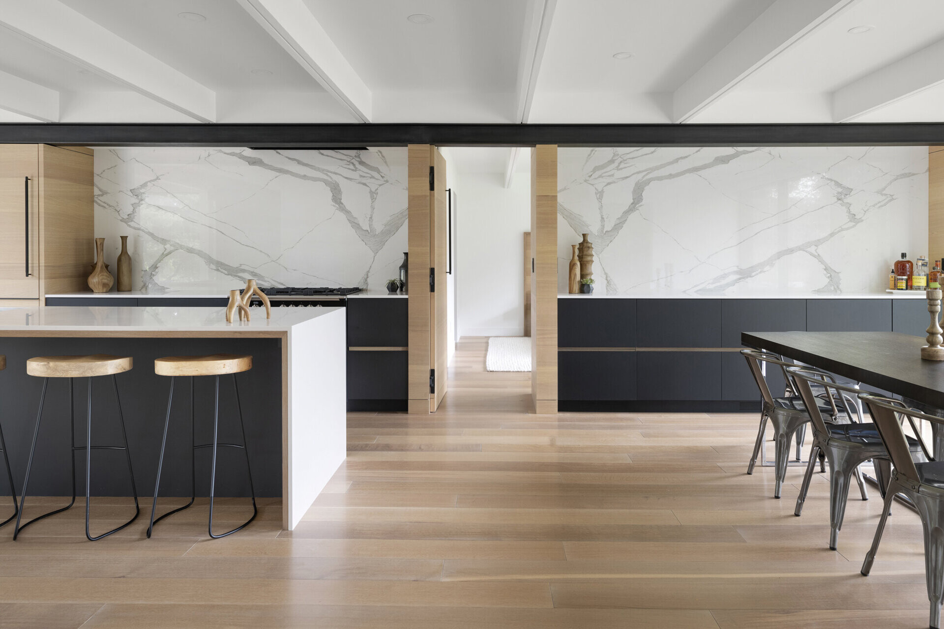El comedor continúa con la paleta de materiales naturales de la casa. Una mesa de comedor de madera rodeada de sillas de color gris oscuro y frente a un botellero negro montado en la pared de ladrillos combina elegancia con calidez.