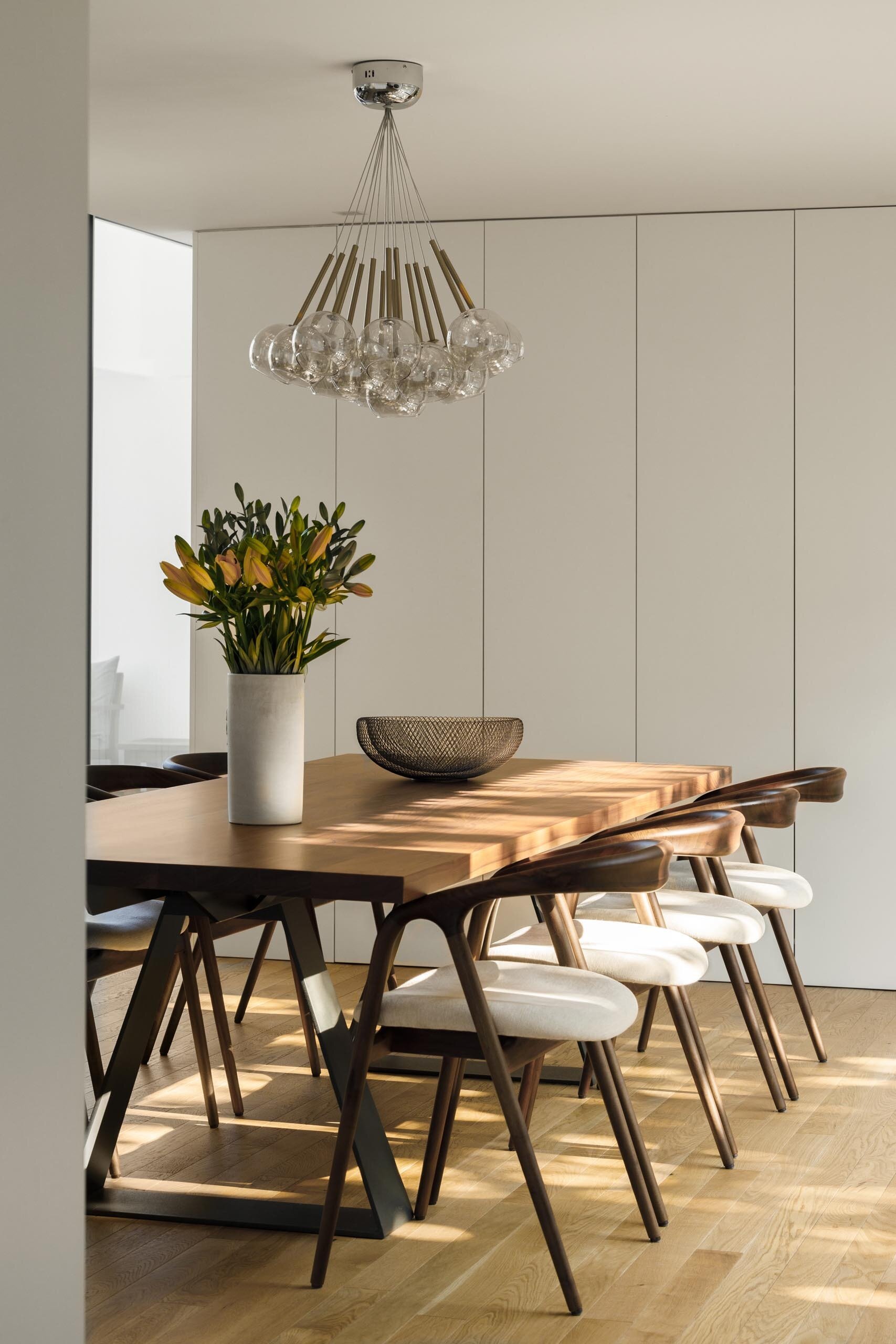 A large wood dining table and soft upholstered chairs create a warm, simple space connected to both kitchen and outdoor areas.