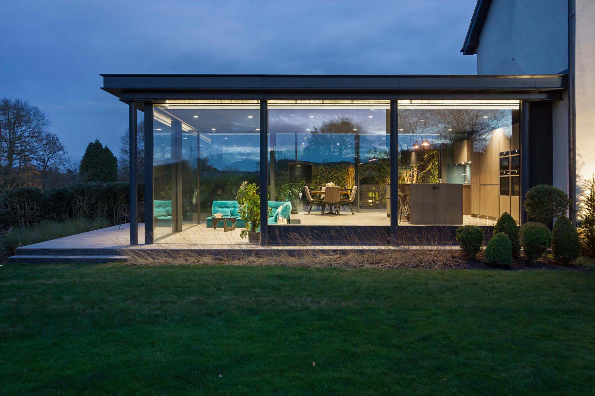 At night, the glass extension glows softly, with the garden becoming a striking backdrop through the glass walls.