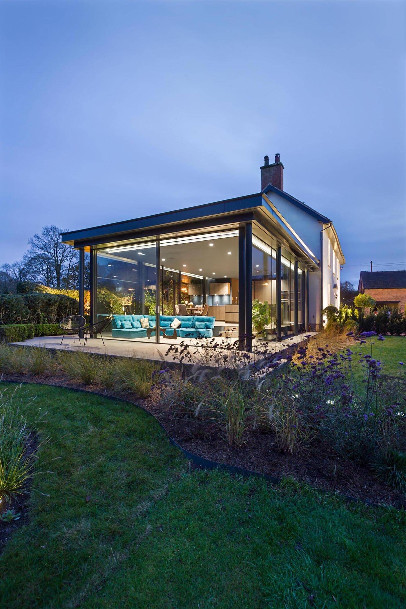 At night, the glass extension glows softly, with the garden becoming a striking backdrop through the glass walls.