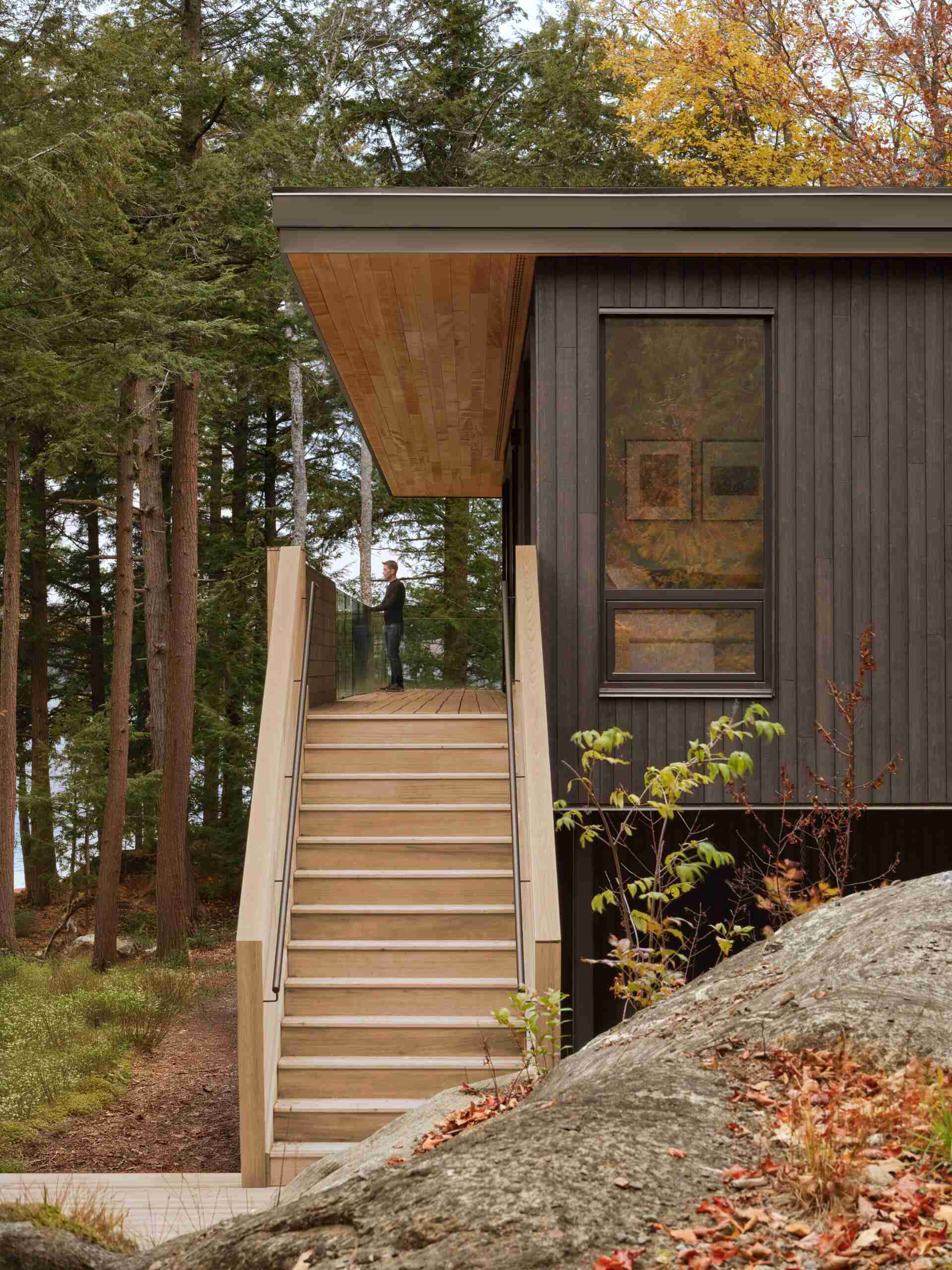 An off-grid cottage set on a forested peninsula, where a bridge leads to an elevated living level that sits lightly among the trees. Clad in greyed cedar and charcoal-stained timber, the exterior blends into the landscape while expansive glazing opens the home to water views on three sides.