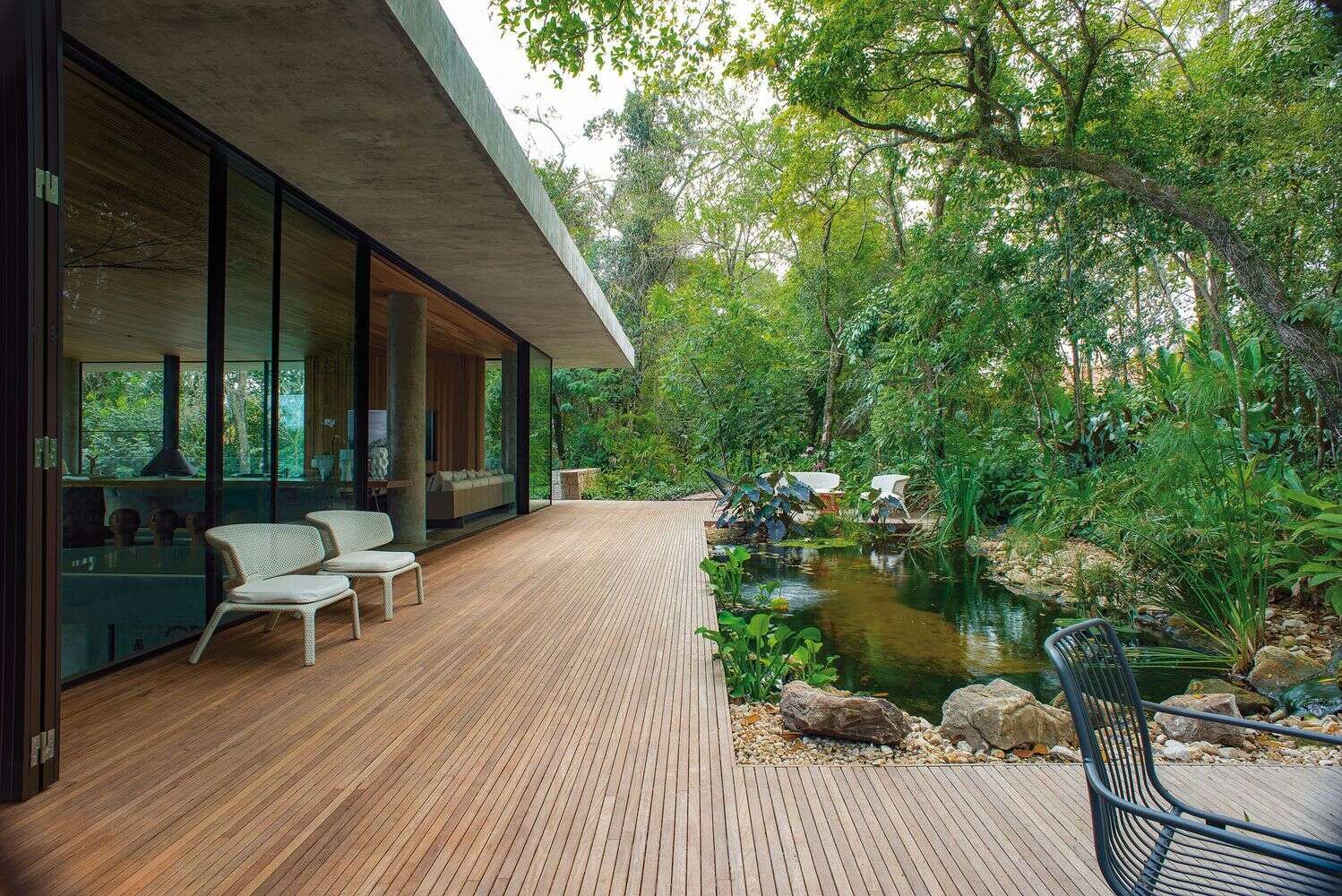 Glass walls open to a wood deck with a natural water feature and tree views, creating a seamless indoor-outdoor connection.
