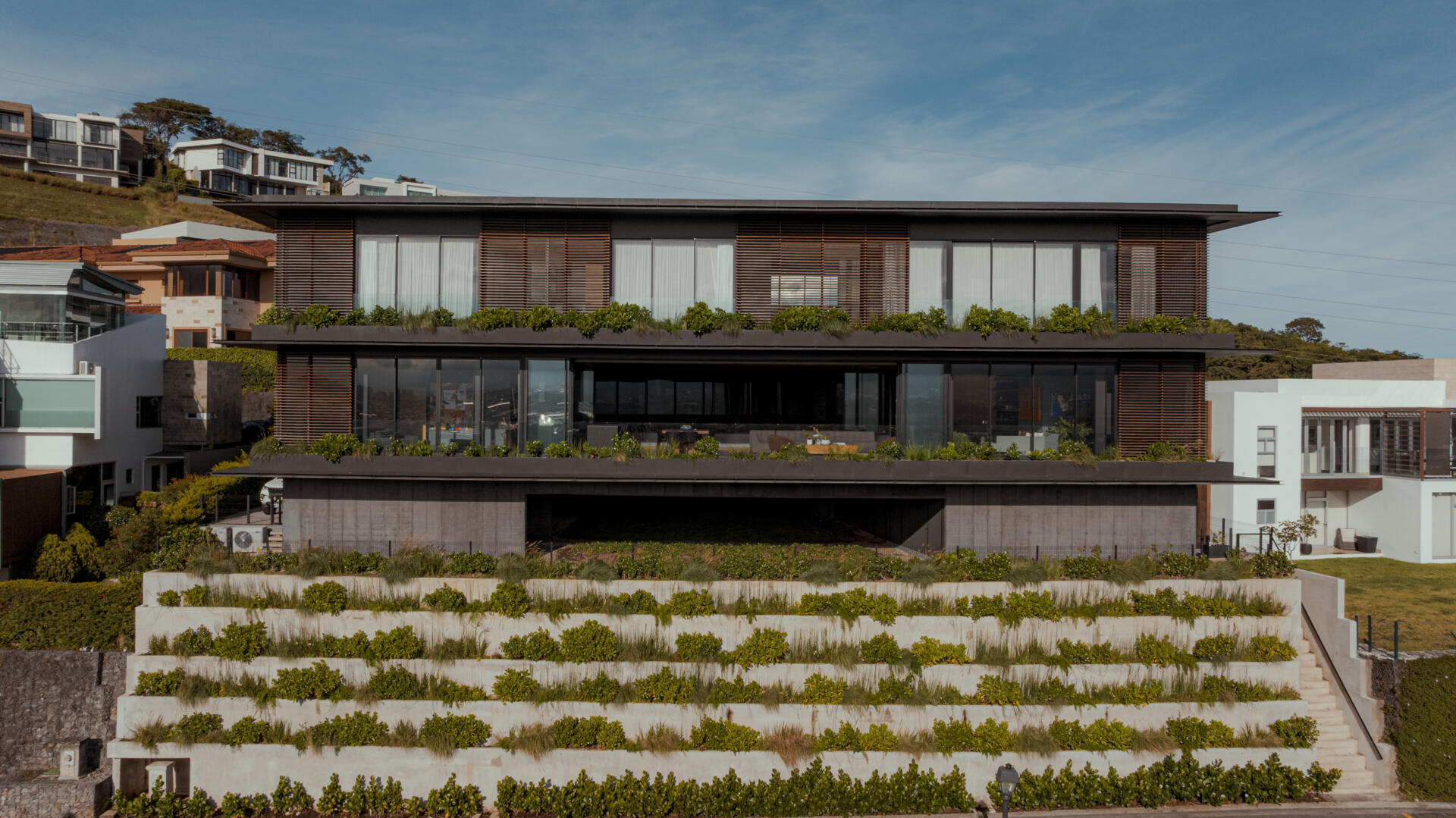 A tiered hillside home in Costa Rica with a central courtyard, open-air spaces, wood slats, and a bridge-like terrace framing expansive valley views.