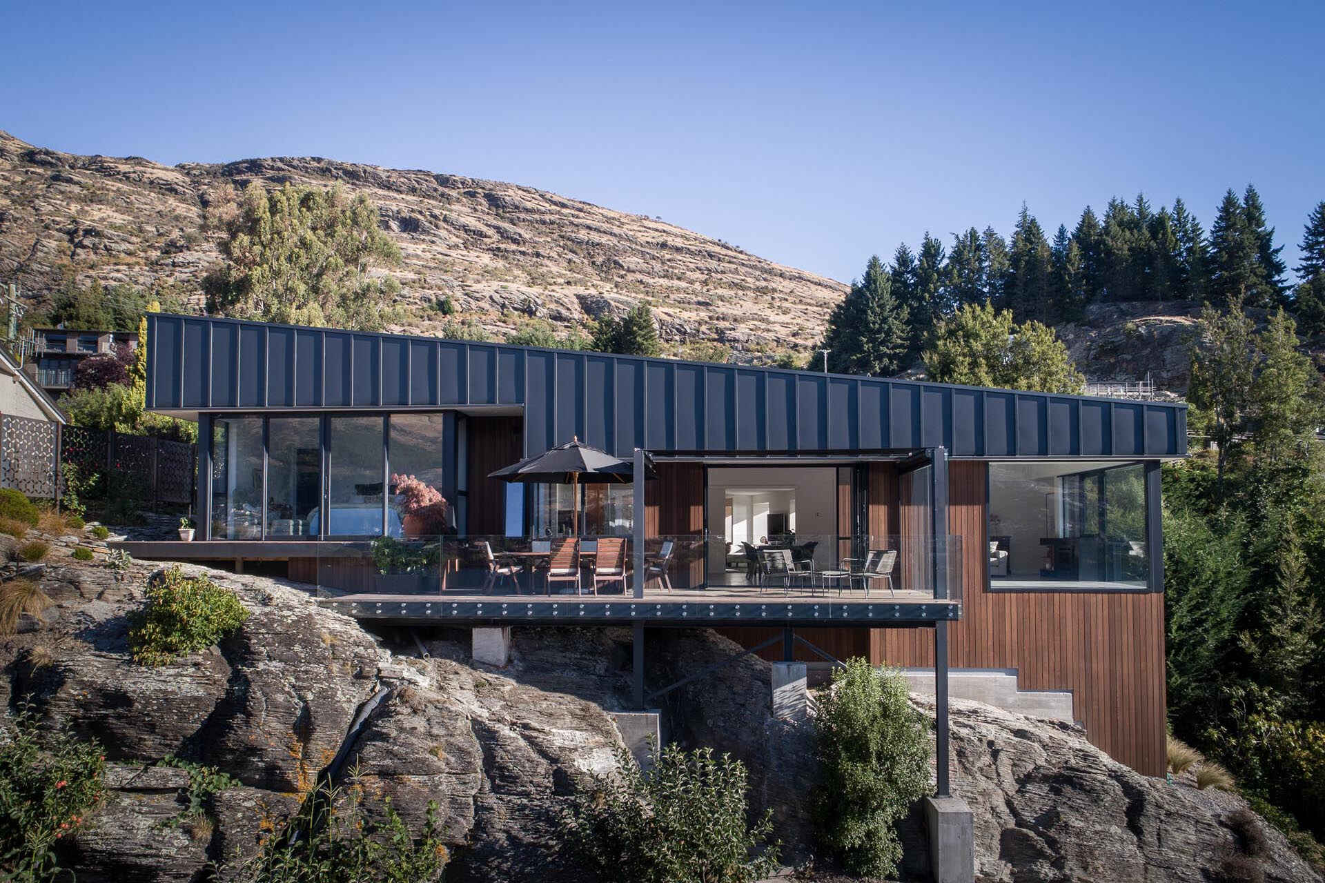 Una casa contemporánea con dos volúmenes en forma de cuña revestidos de metal negro mate y escalones de madera que descienden por un empinado sitio de Queenstown mientras enmarcan las vistas del lago Wakatipu.