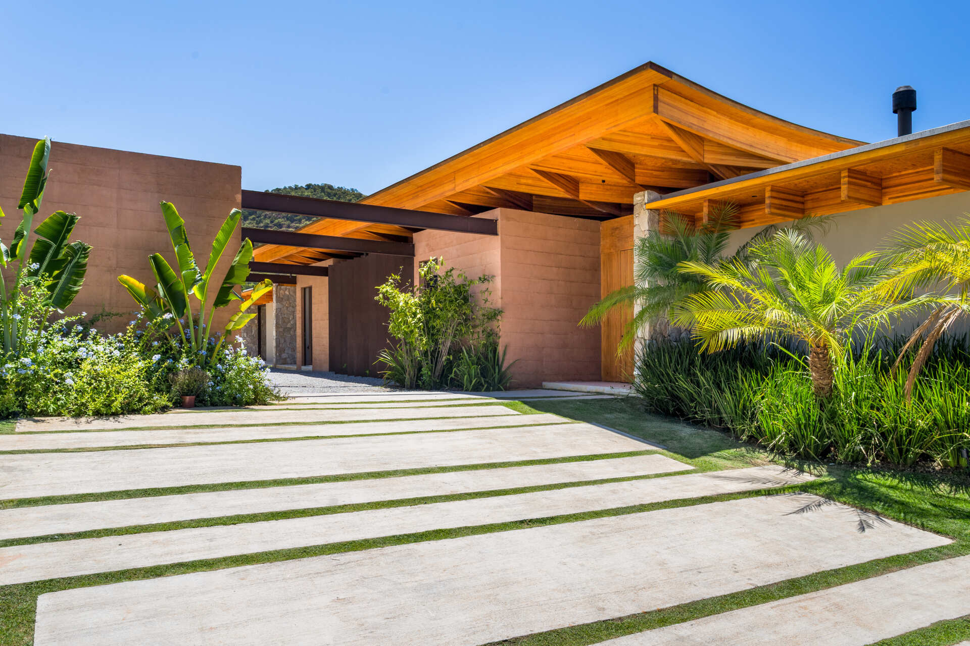 Concrete slabs set into grass lead through dense vegetation toward stone and terracotta volumes. The house unfolds gradually, inviting discovery across the plateau with views of greenery and a nearby lake.