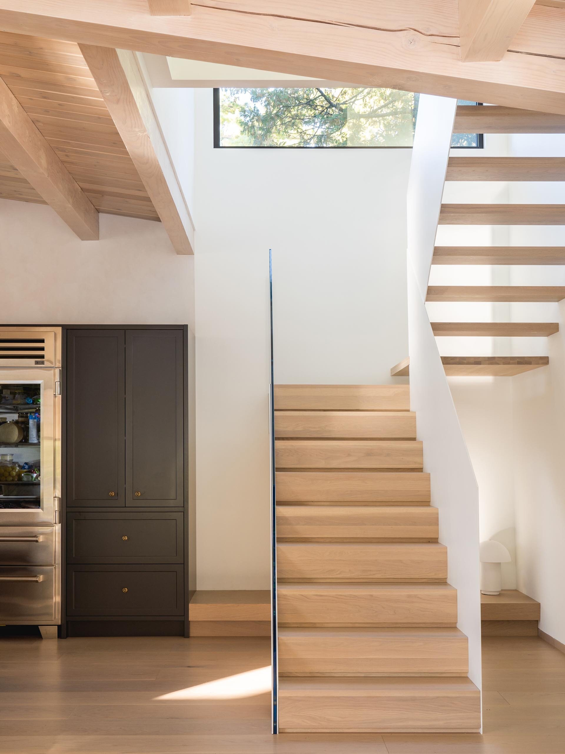 A staircase with a white metal frame, wood treads, and a glass railing allows daylight to pass through the interior while leading to the new upper level.