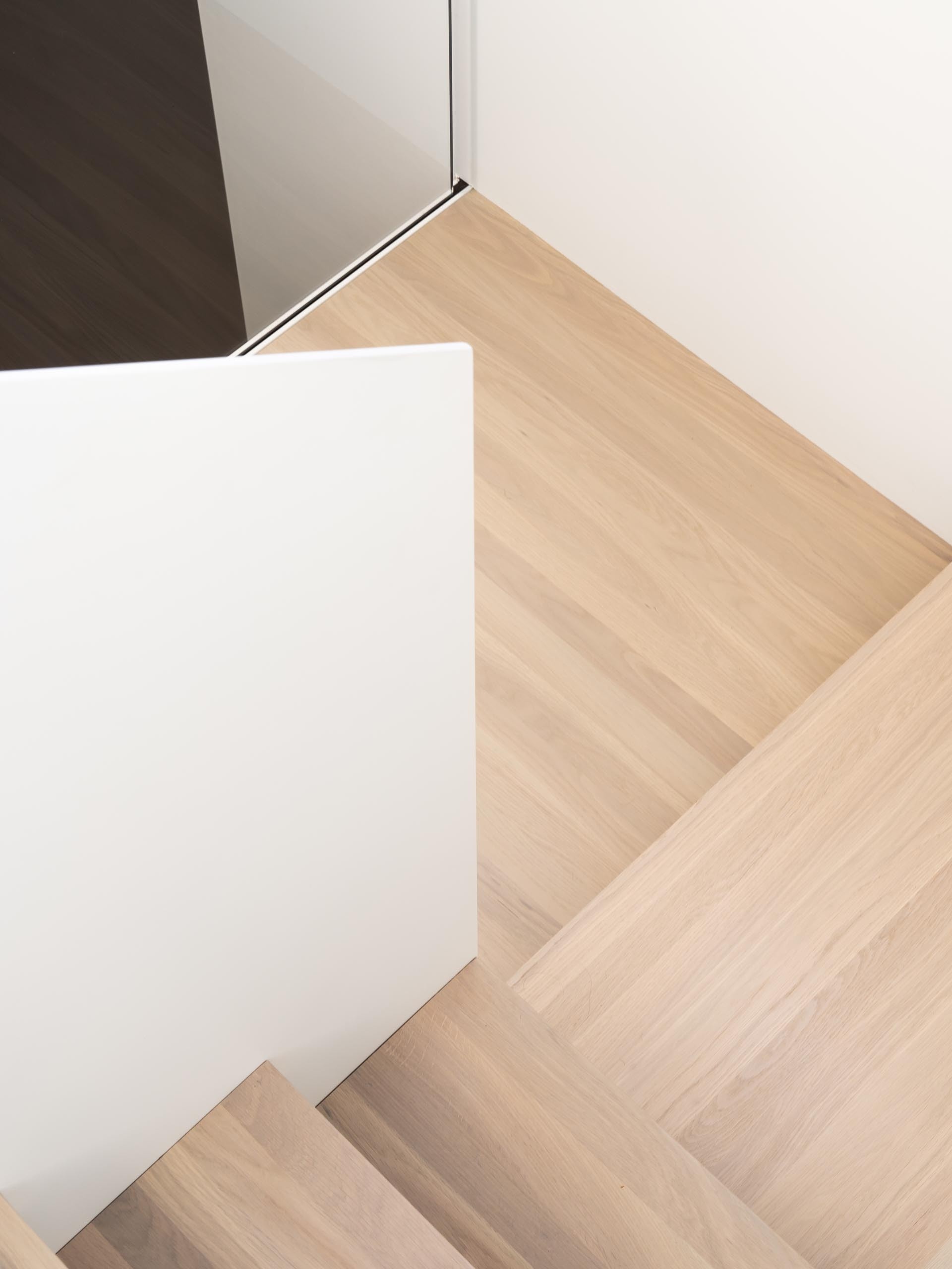 A staircase with a white metal frame, wood treads, and a glass railing allows daylight to pass through the interior while leading to the new upper level.